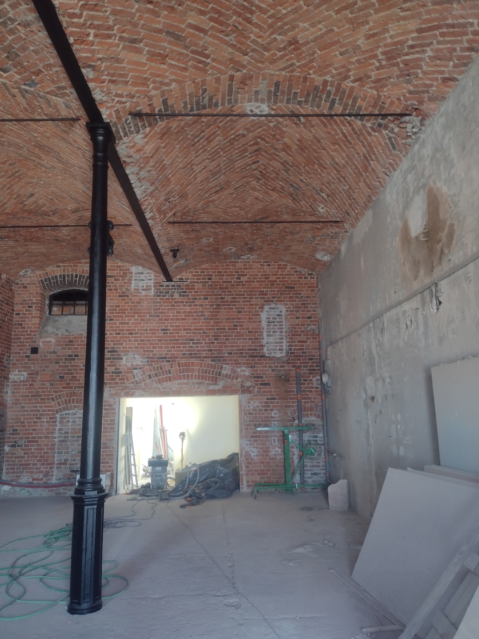 Wnętrze w trakcie renowacji: sklepione ceglane sufity, czarna metalowa kolumna, ściany z surowego betonu i cegły, widoczne narzędzia i materiały budowlane.