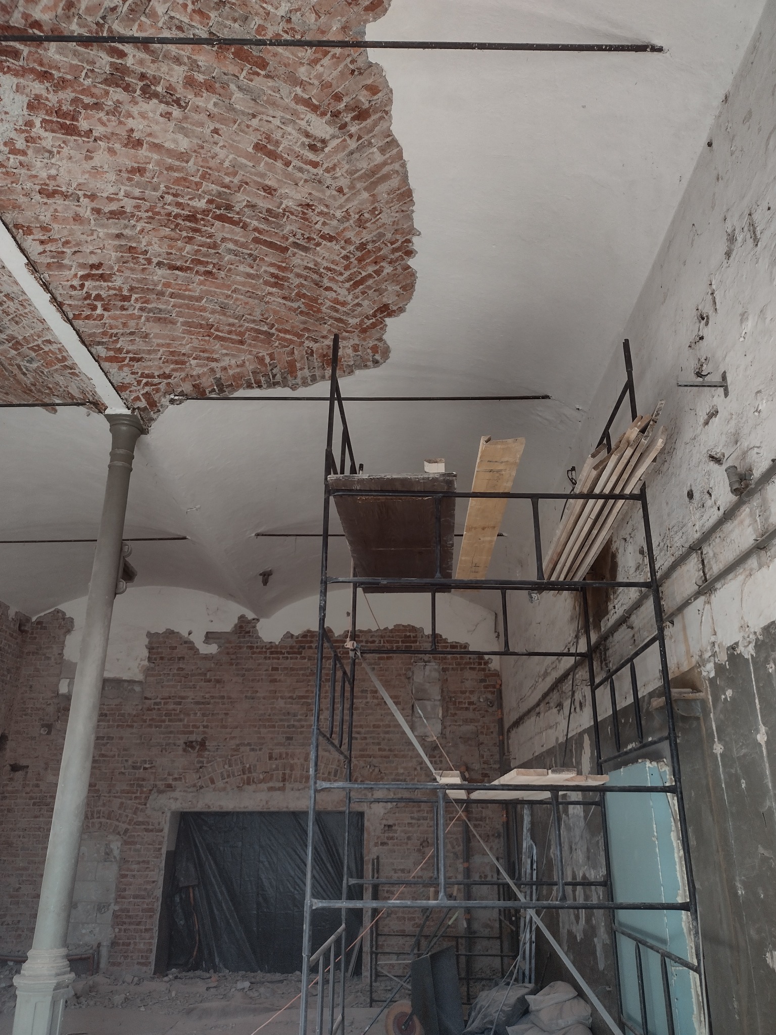 Wnętrze budynku w trakcie prac remontowych: odsłonięta cegła na sklepieniu, rusztowanie z deskami, kolumna i gruz na podłodze.