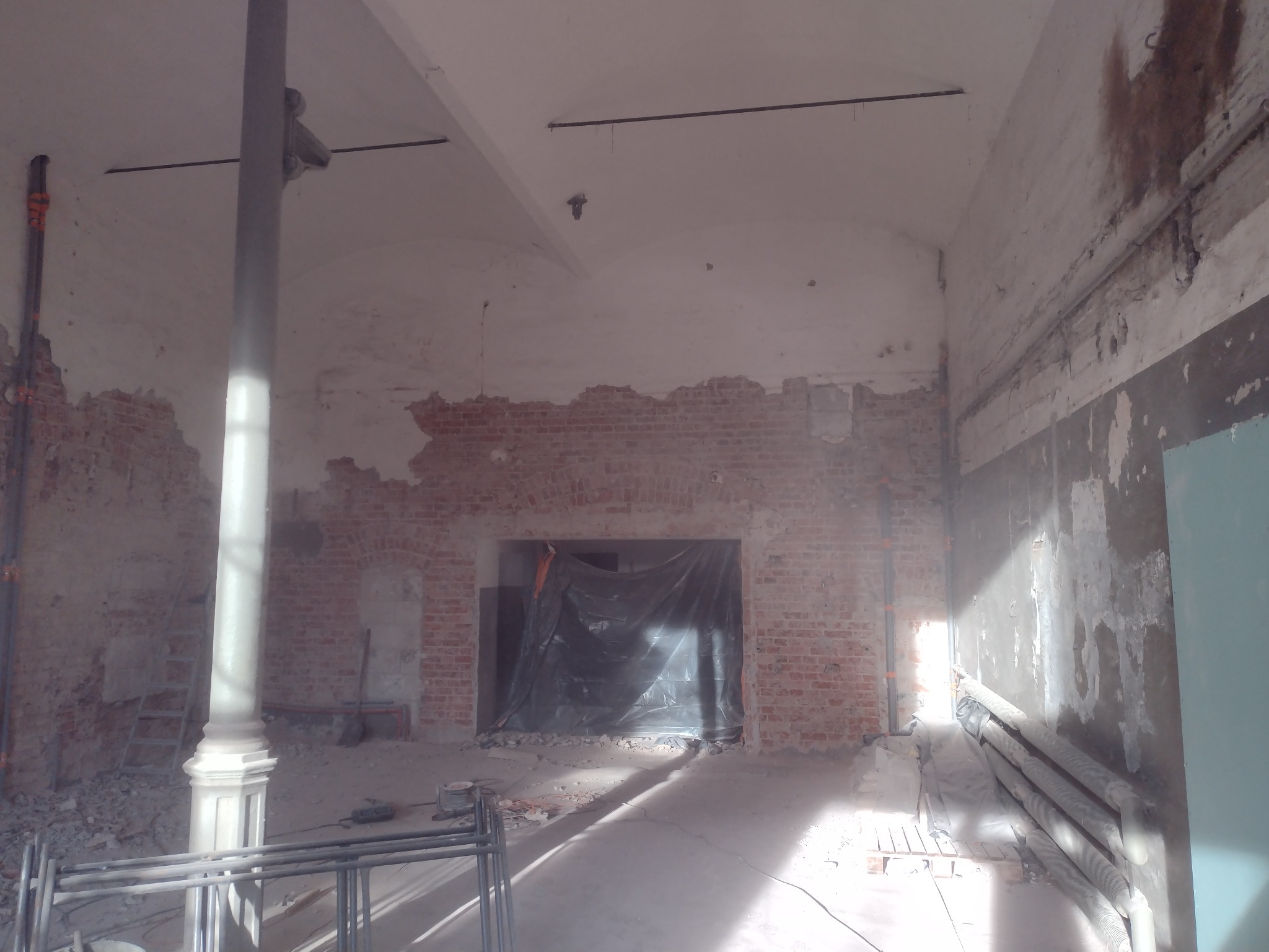 Wnętrze w trakcie rozbiórki: fragmenty ceglanej ściany, gruz, zabezpieczony otwór folią, kolumna podtrzymująca, widoczne instalacje.