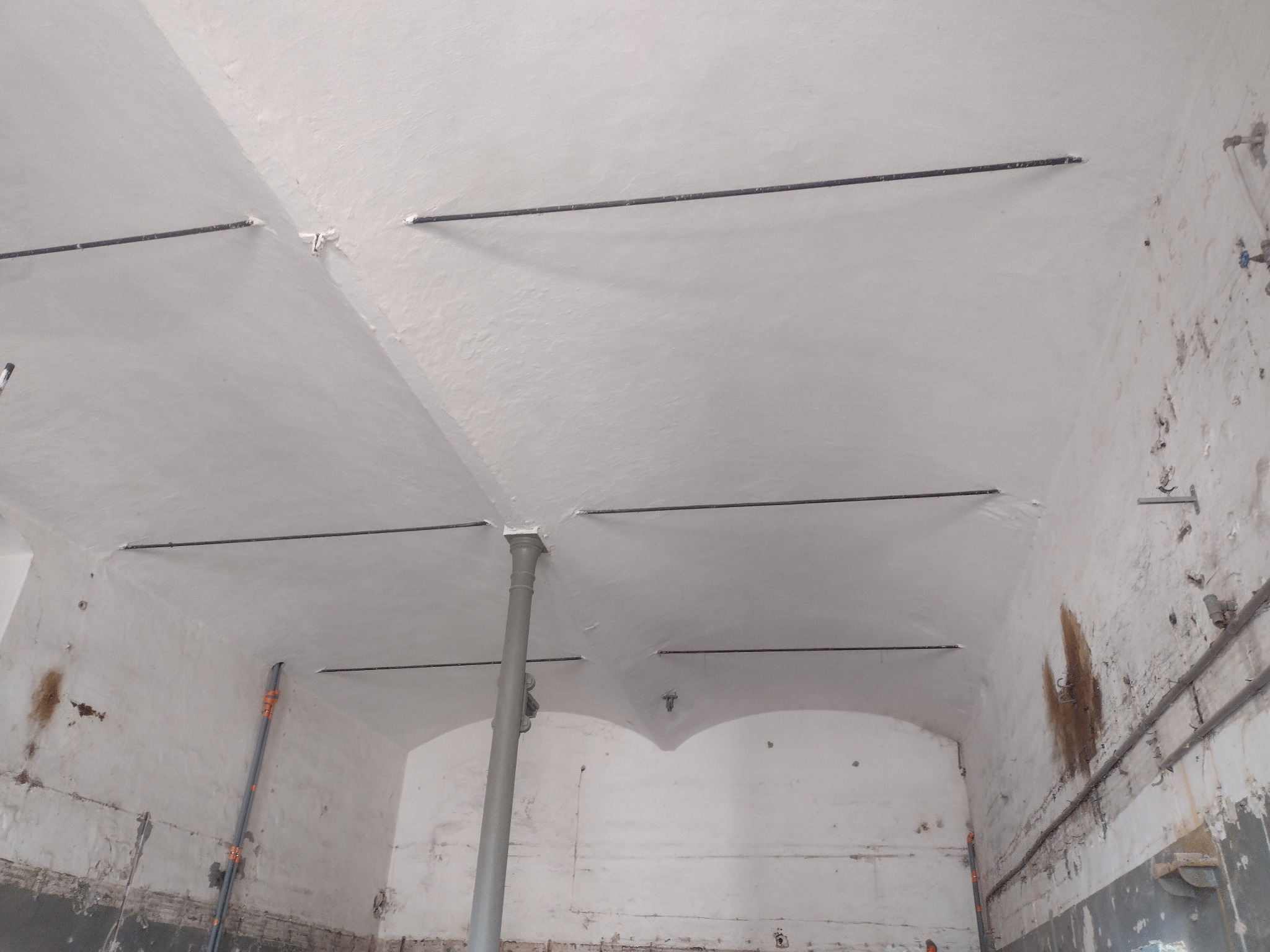 Wnętrze pomieszczenia z łukowatym sufitem, pomalowanym na biało, z widocznymi instalacjami pod sufitem i śladami wilgoci na ścianach.