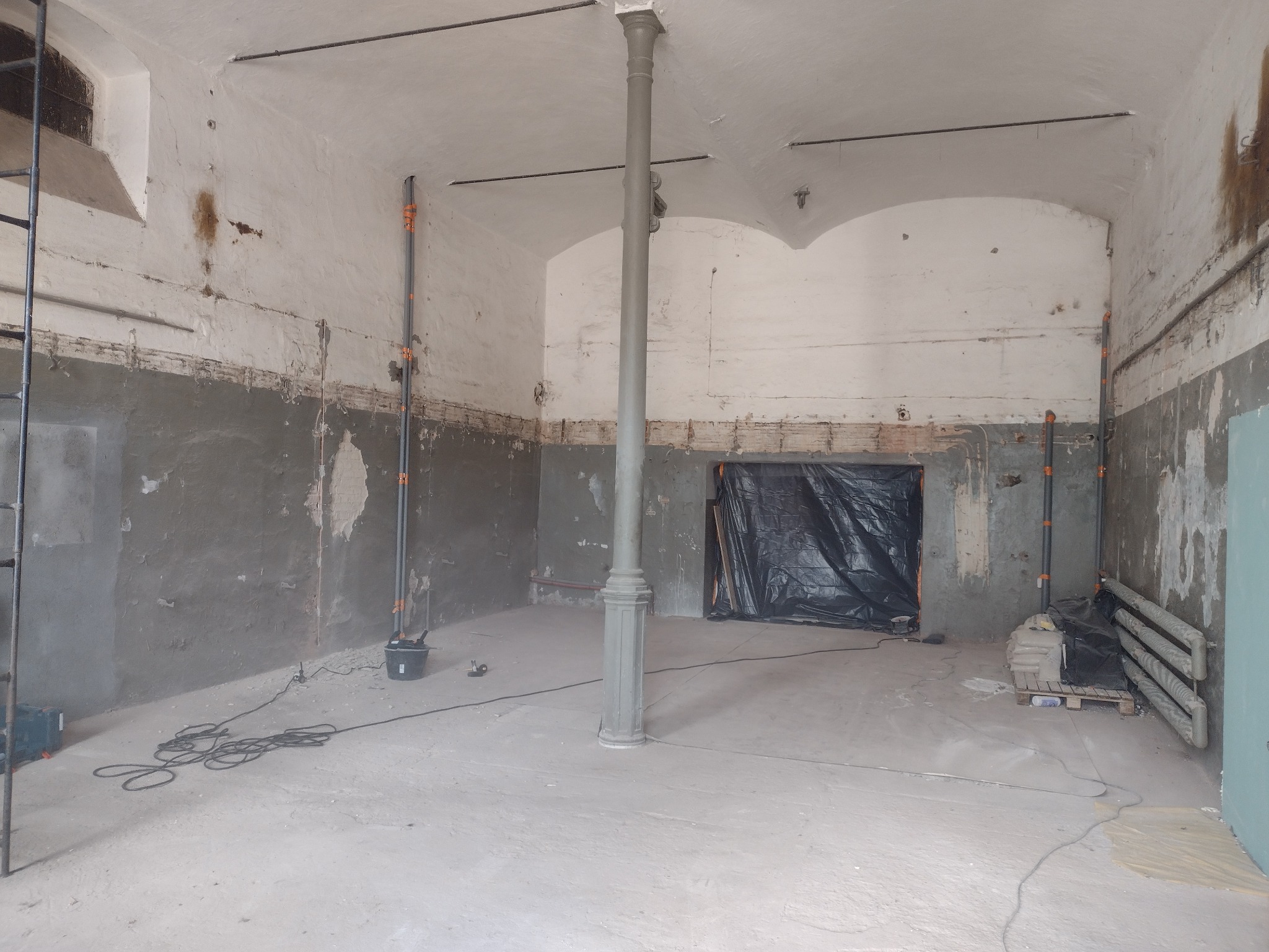 Wnętrze pomieszczenia w trakcie prac przygotowawczych do remontu, widoczne odsłonięte fragmenty ścian, zabezpieczone folią otwory, pionowa kolumna konstrukcyjna, oraz narzędzia i materiały budowlane.