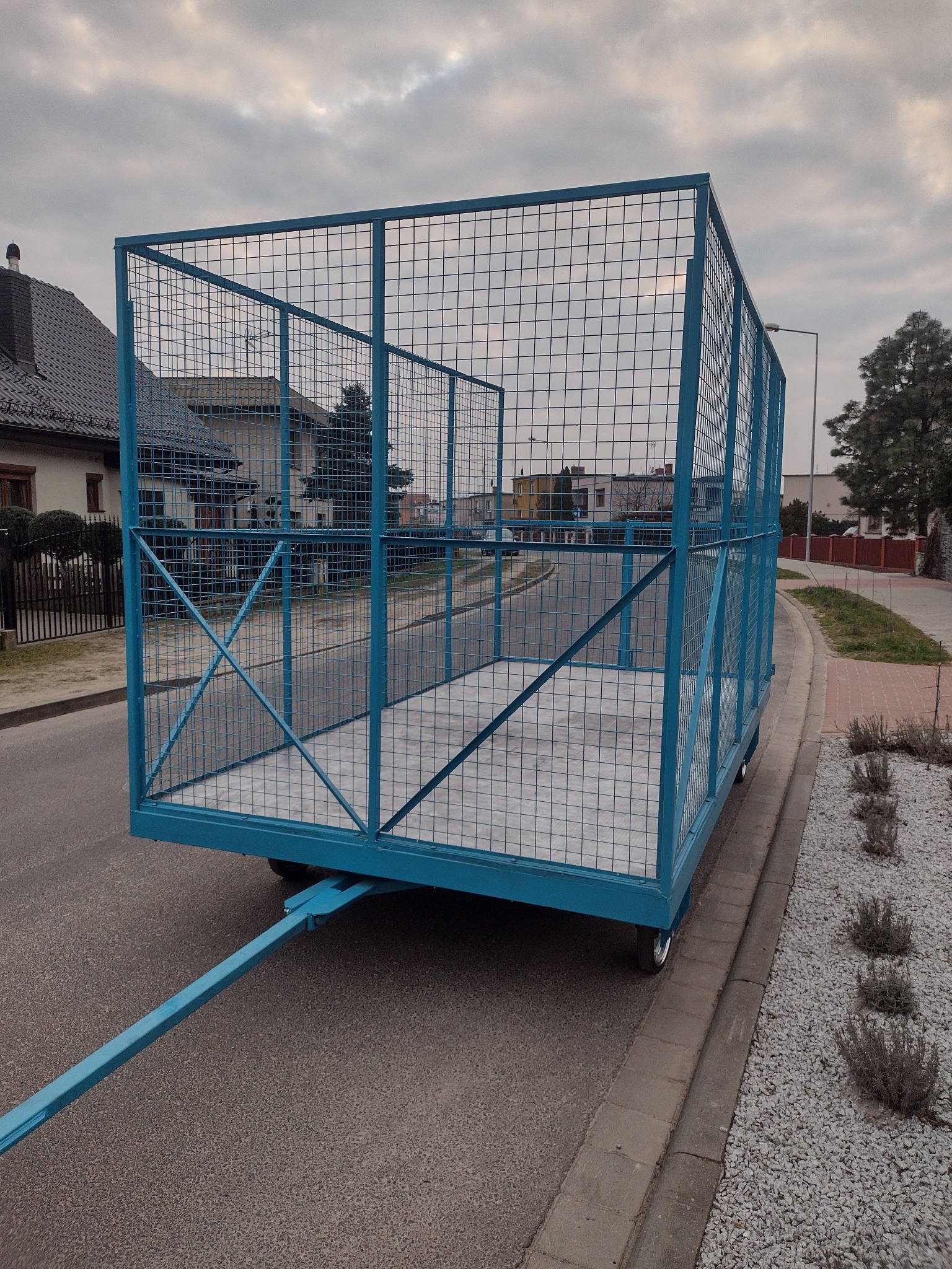 Błękitny kontener siatkowy na kółkach z dyszlem, stojący na ulicy w zabudowie jednorodzinnej, prawdopodobnie przeznaczony do transportu gałęzi lub innych odpadów po wycince drzew.