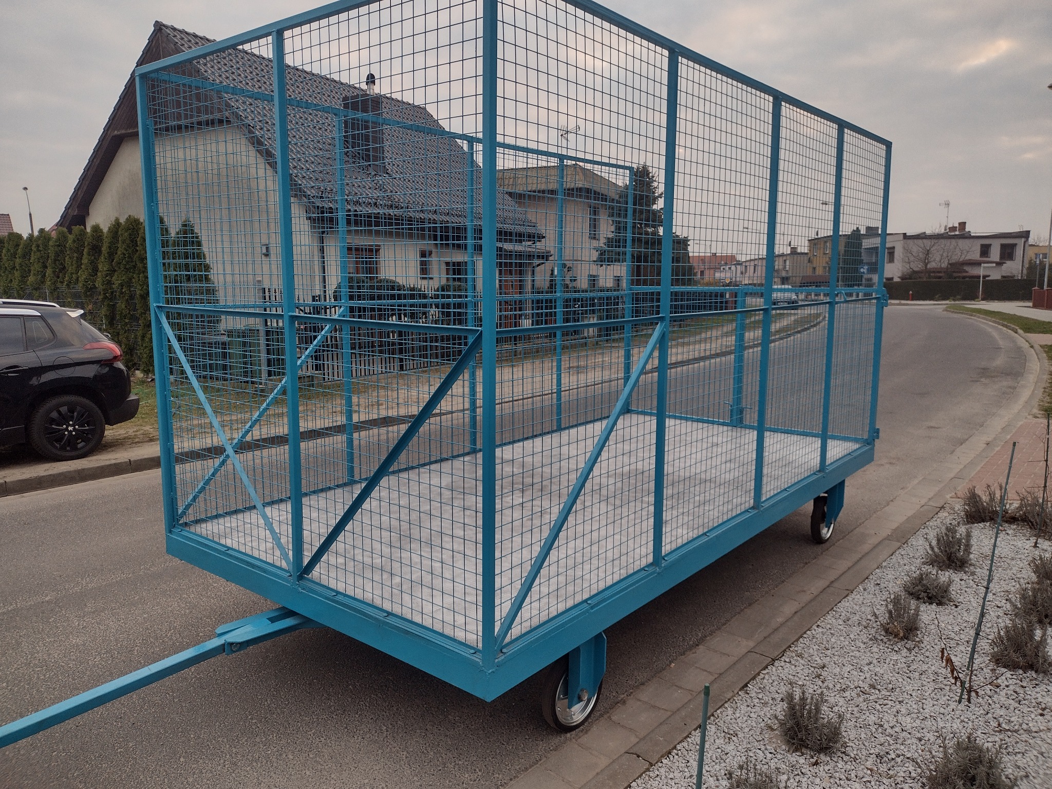 Niebieski, metalowy wózek z siatką na kółkach, zaparkowany na ulicy w Poznaniu, z domami w tle.