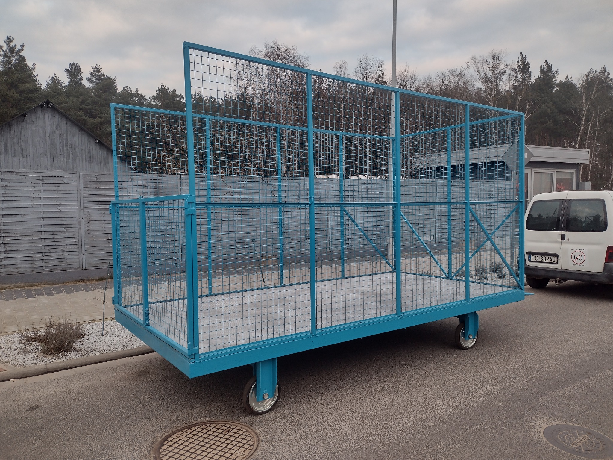 Błękitny, metalowy kontener siatkowy na kółkach, stojący na asfalcie w pobliżu zabudowań i zaparkowanego białego samochodu dostawczego.