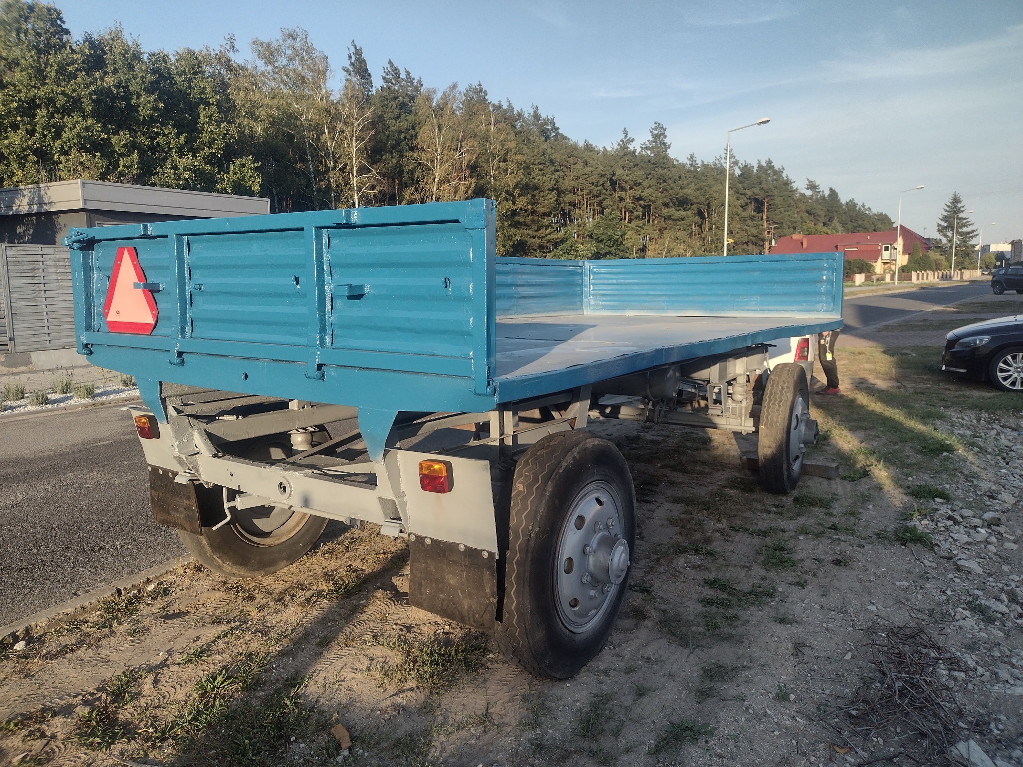 Niebieska, odnowiona przyczepa samochodowa z trójkątnym znakiem ostrzegawczym, stojąca na ziemi z widocznymi śladami opon i zaroślami, na tle drzew i budynków.