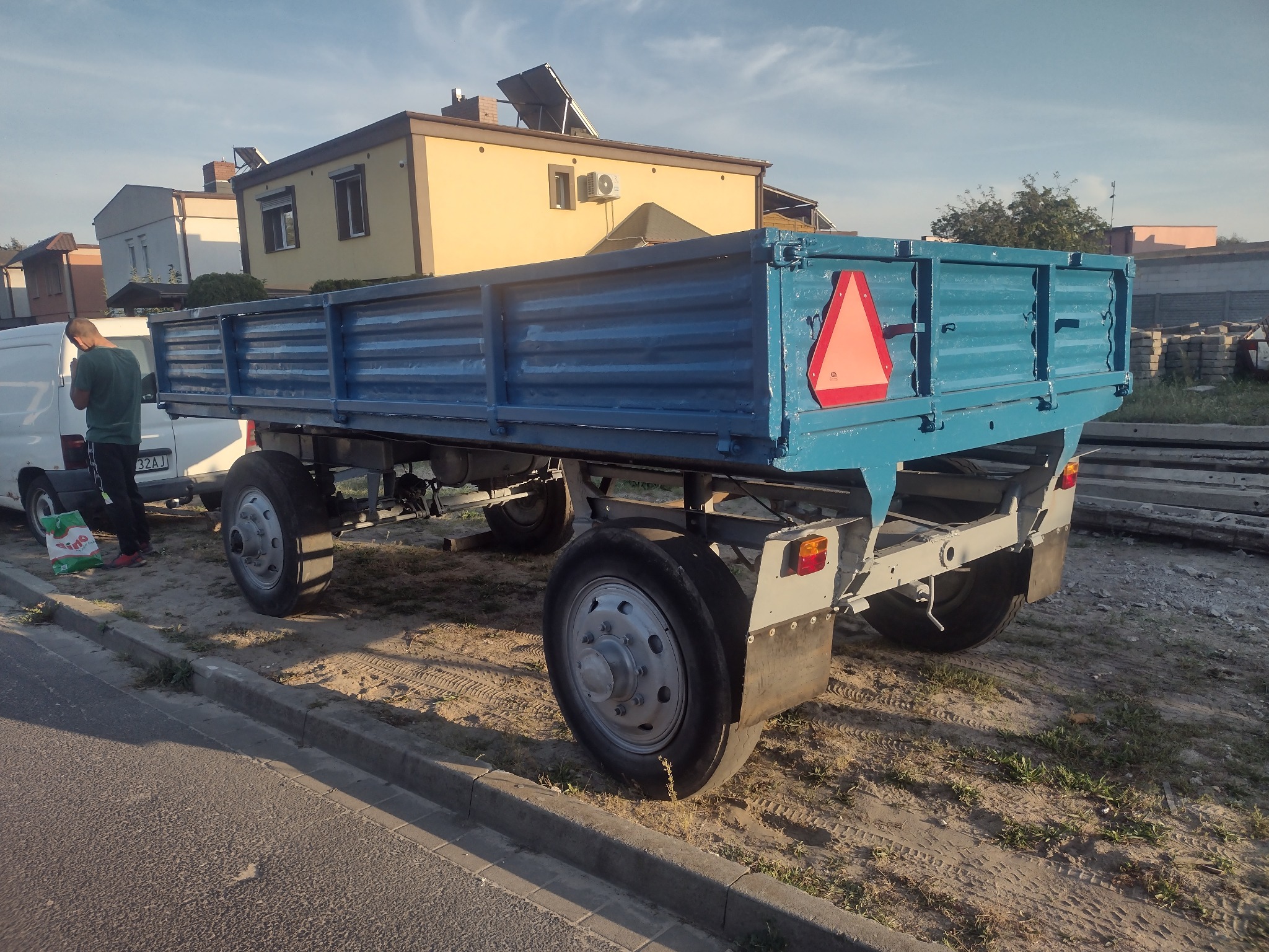 Błękitna przyczepa rolnicza z czerwoną tablicą ostrzegawczą, zaparkowana na piaszczystym podłożu przy krawężniku, z widocznym fragmentem białego busa i budynków w tle.