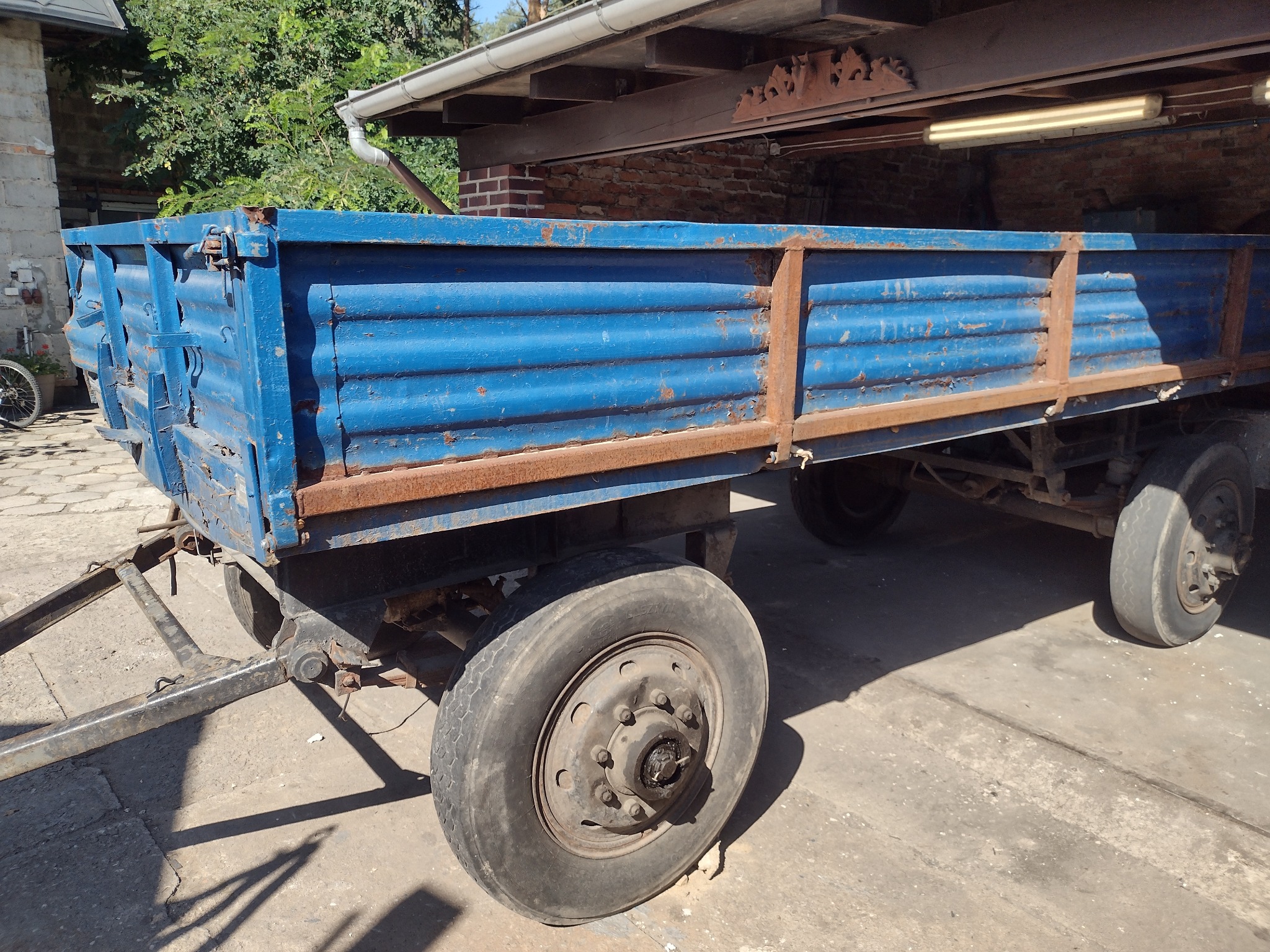 Przyczepa rolnicza z niebieską skrzynią ładunkową i rdzawymi elementami metalowymi, stojąca na betonowym podłożu w cieniu budynku.