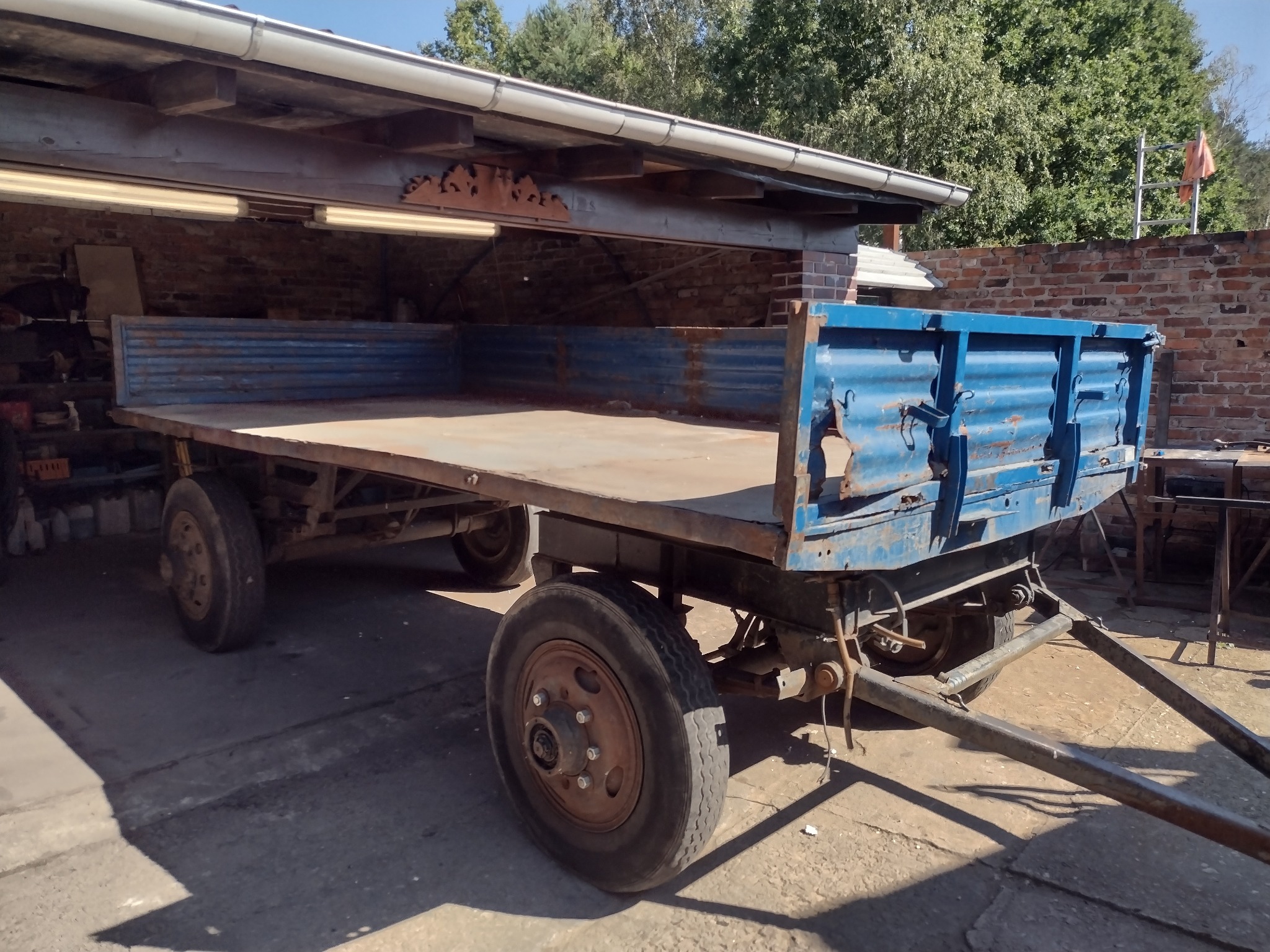 Stara, niebieska przyczepa rolnicza z płaską platformą, zaparkowana pod zadaszeniem z ceglanym murem w tle.