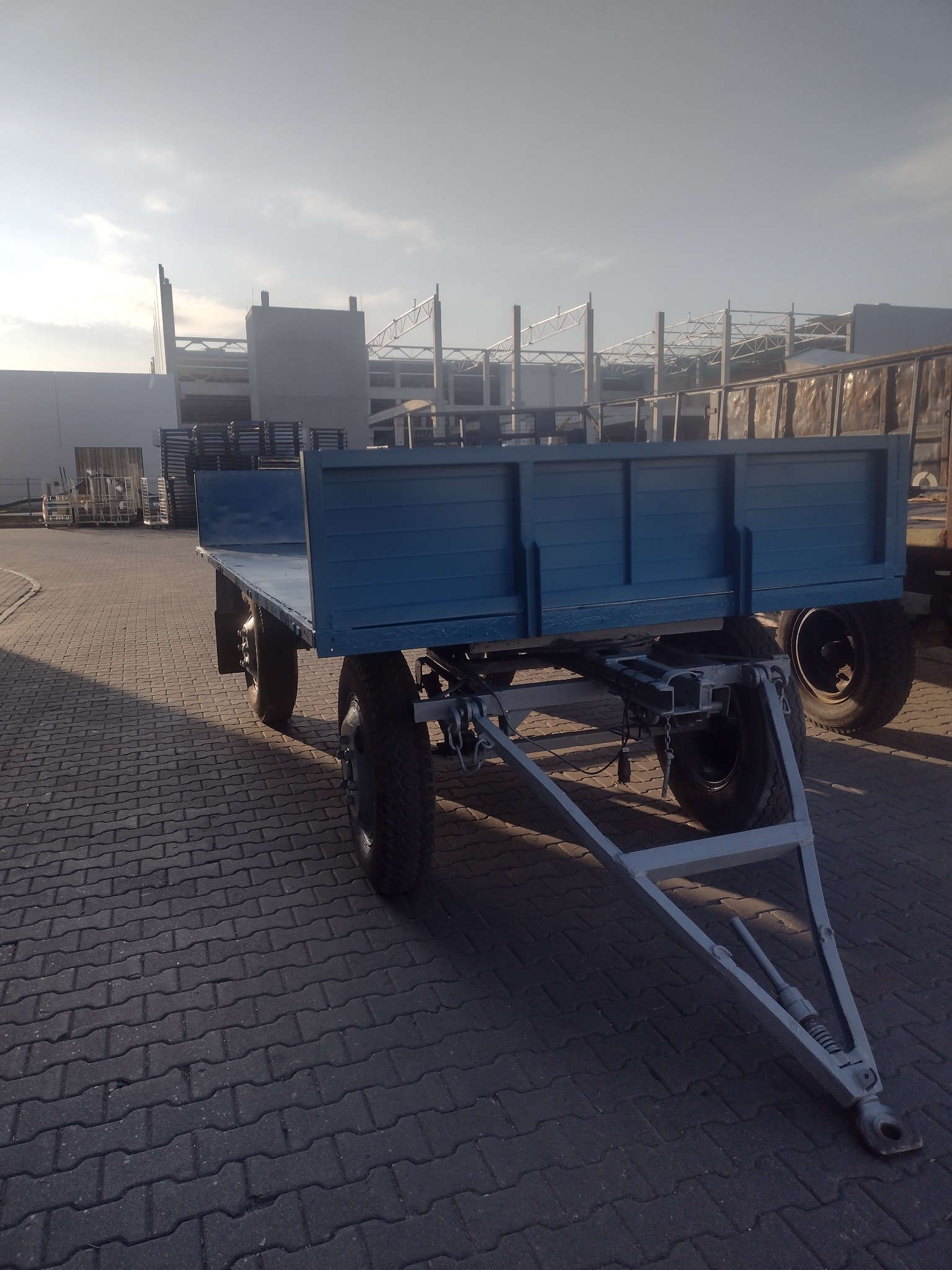 Niebieska przyczepa rolnicza z szarym podwoziem, stojąca na kostce brukowej, w tle budynek w trakcie budowy z elementami stalowej konstrukcji.