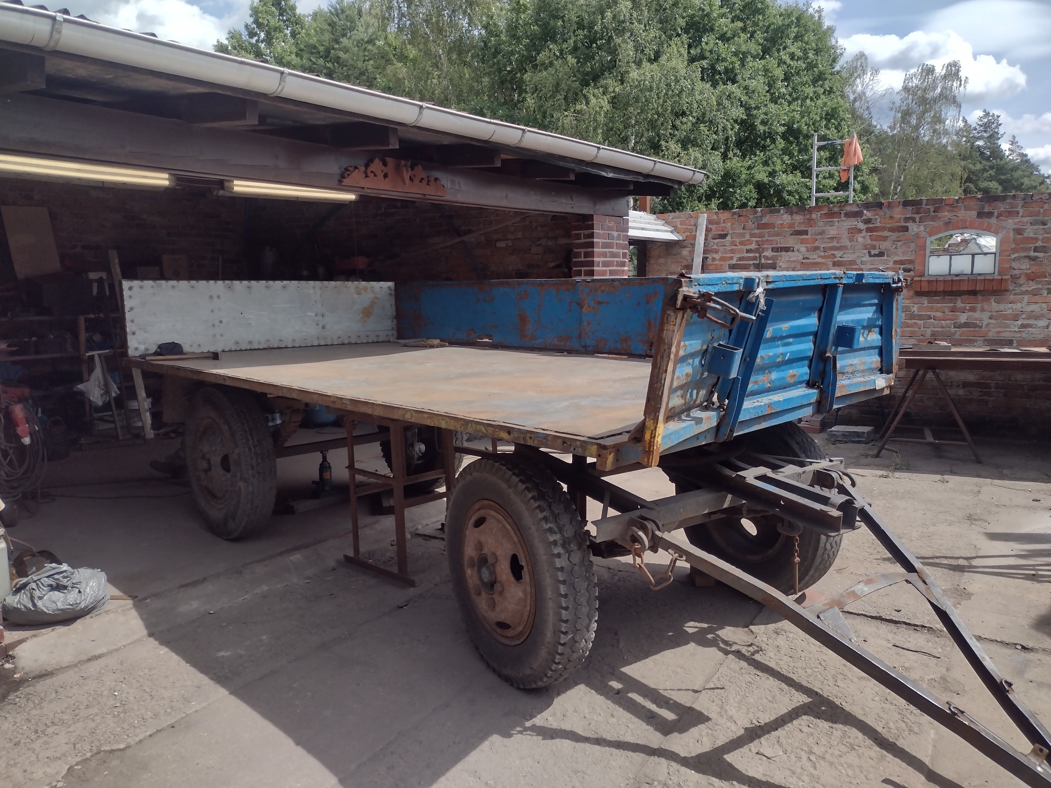 Stara, zardzewiała przyczepa rolnicza z metalową platformą i niebieską skrzynią, stojąca w zadaszonym garażu z ceglanymi ścianami i oświetleniem jarzeniowym.