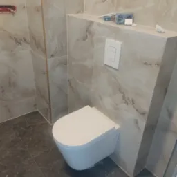 Wisząca toaleta z zabudową geberitową w łazience wykończonej marmurkowymi płytkami; widoczne niedokończone prace elektryczne na półce.