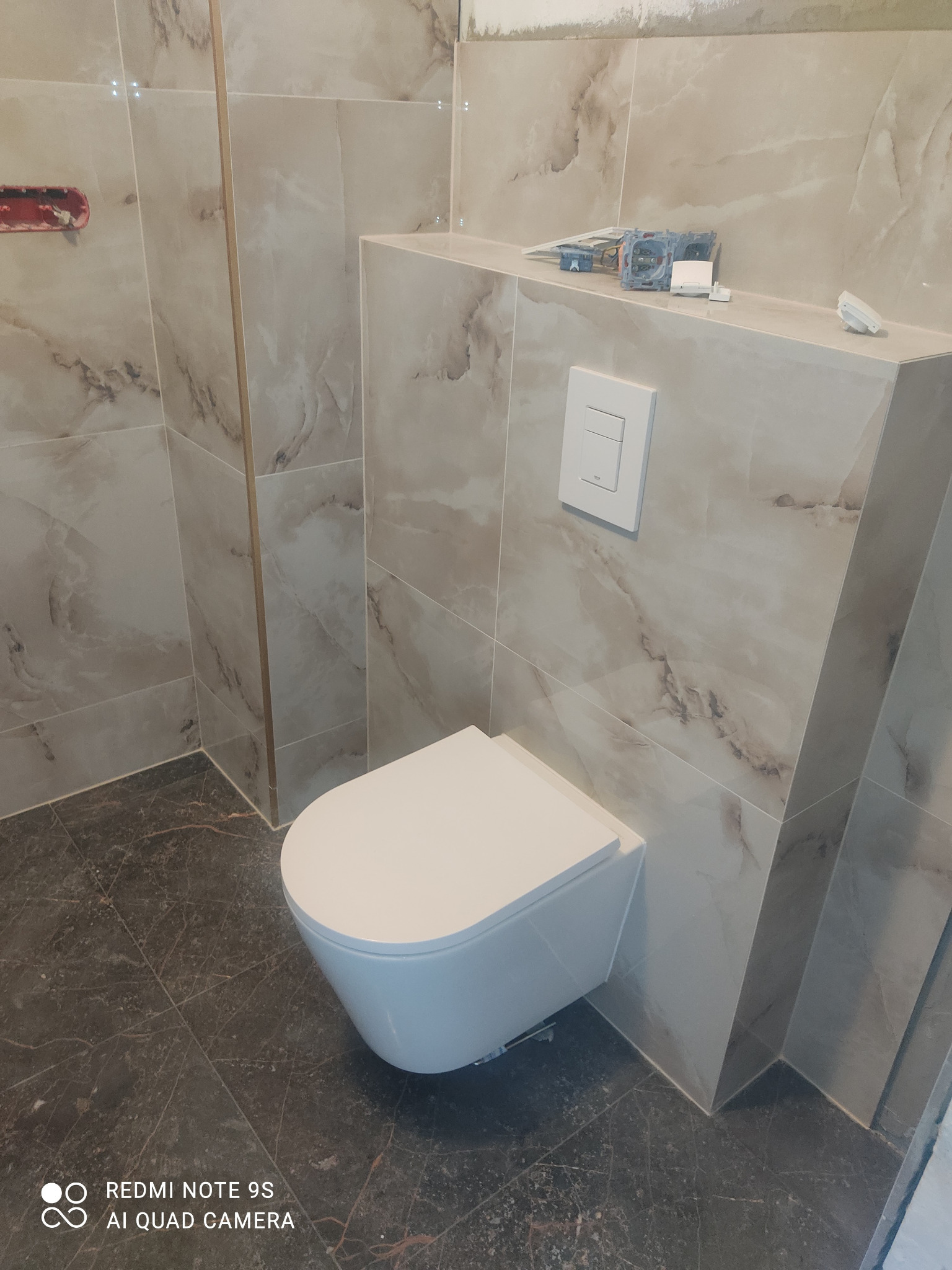 Wisząca toaleta z zabudową geberitową w łazience wykończonej marmurkowymi płytkami; widoczne niedokończone prace elektryczne na półce.