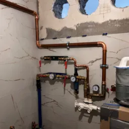 Miedziana instalacja wodna z zaworami, manometrami i filtrem, zamontowana na ścianie z marmurowymi płytkami, widoczne otwory w ścianie przygotowane do dalszych prac wykończeniowych.