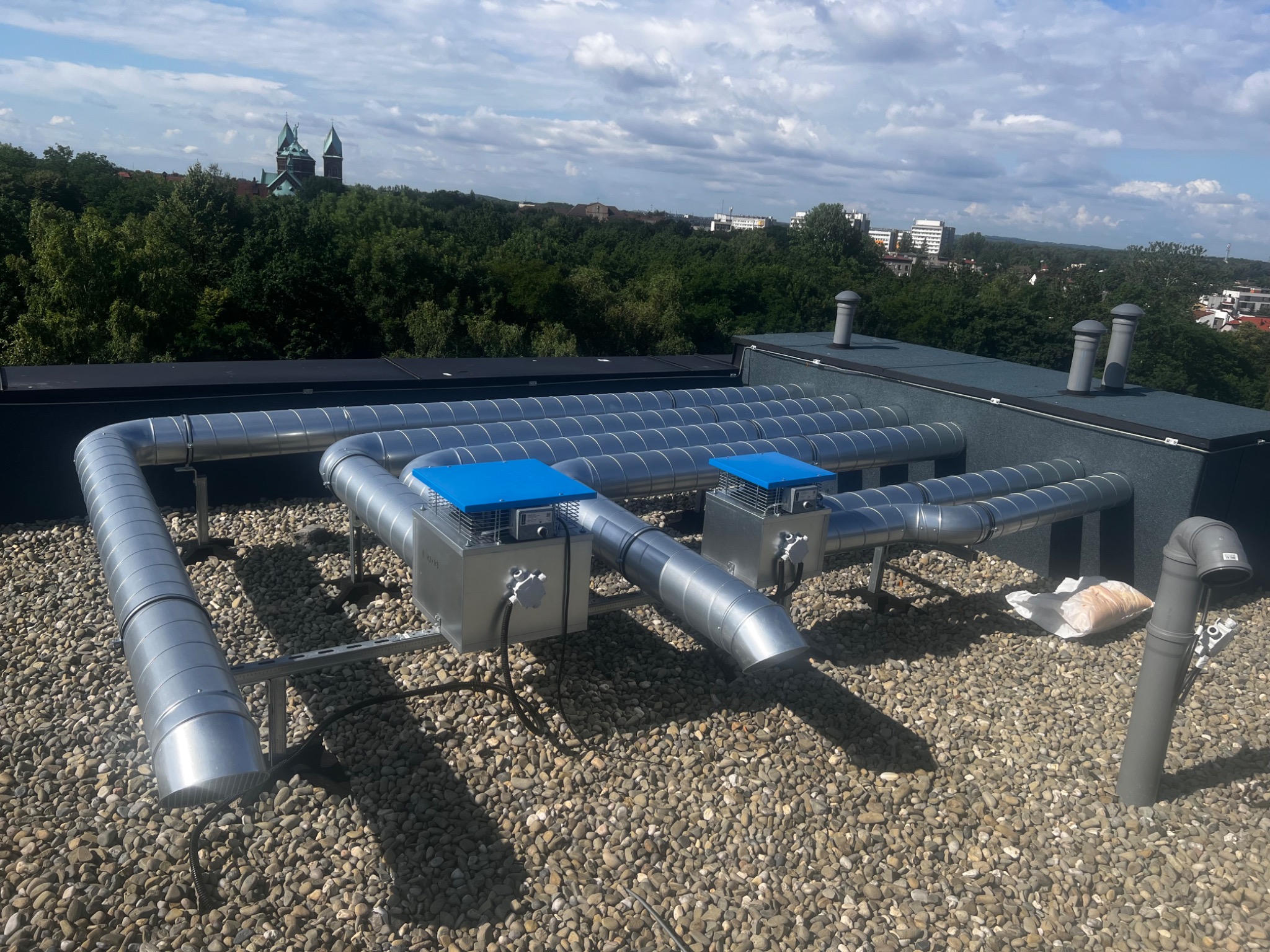 Instalacja wentylacyjna z metalowych rur na dachu budynku z widokiem na miasto i kościół w tle.
