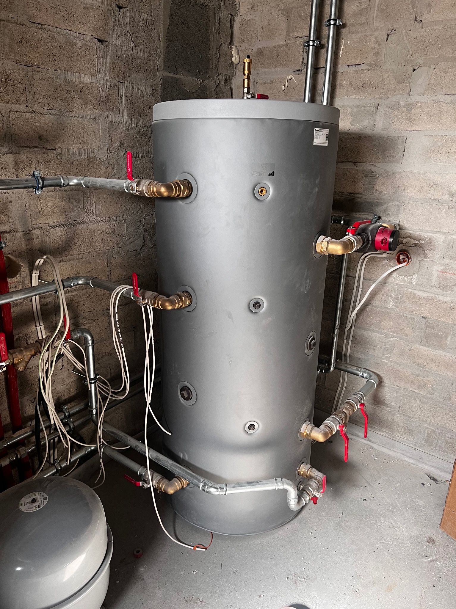 Szary, cylindryczny zasobnik ciepłej wody z licznymi przyłączami rur miedzianych i zaworami z czerwonymi uchwytami na tle surowej ściany z cegły.