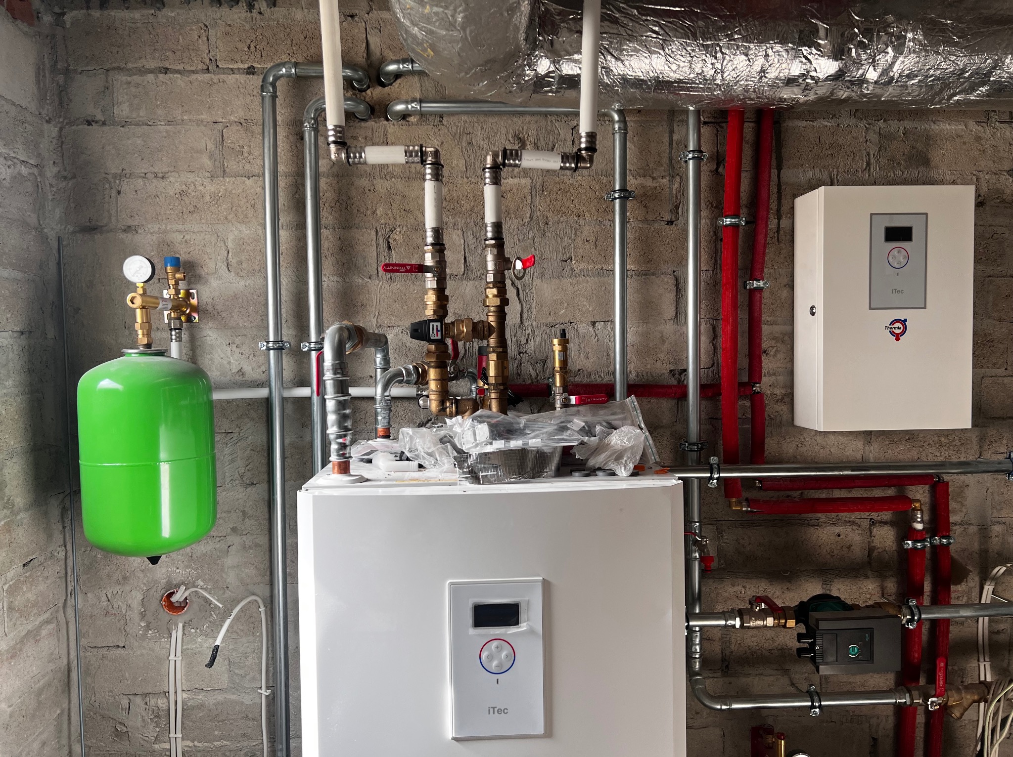 Nowa instalacja grzewcza z zielonym zbiornikiem wyrównawczym, białym piecem i systemem rur na tle surowej ściany z cegły.