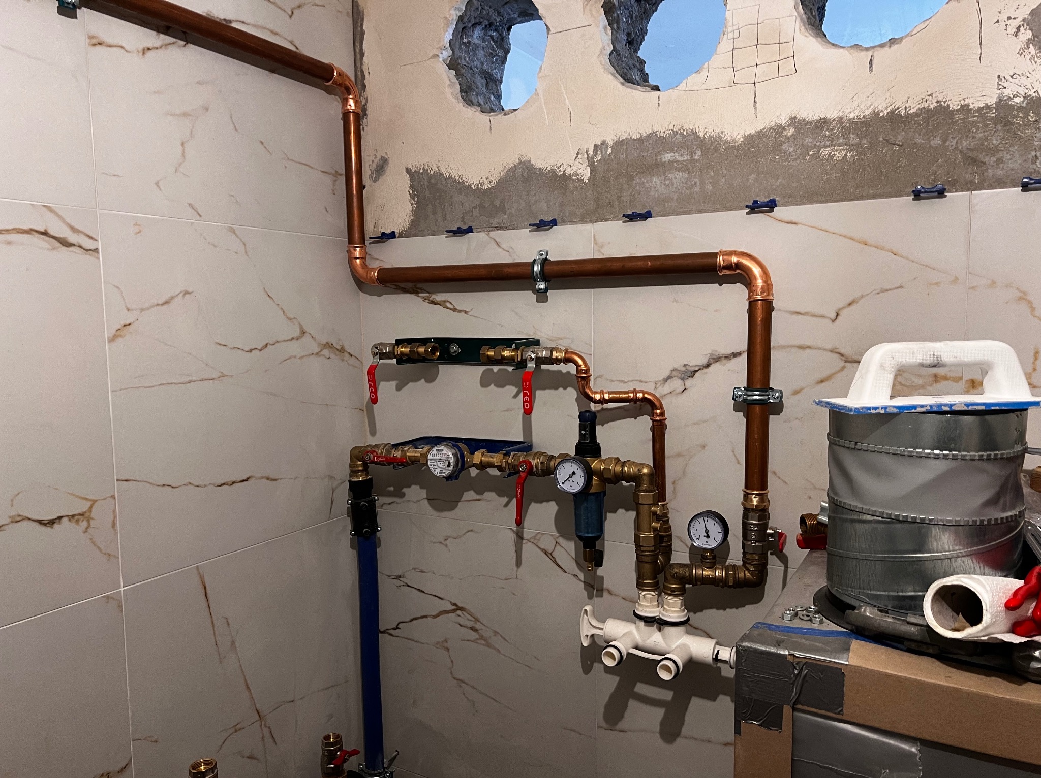 Miedziana instalacja wodna z zaworami, manometrami i filtrem, zamontowana na ścianie z marmurowymi płytkami, widoczne otwory w ścianie przygotowane do dalszych prac wykończeniowych.