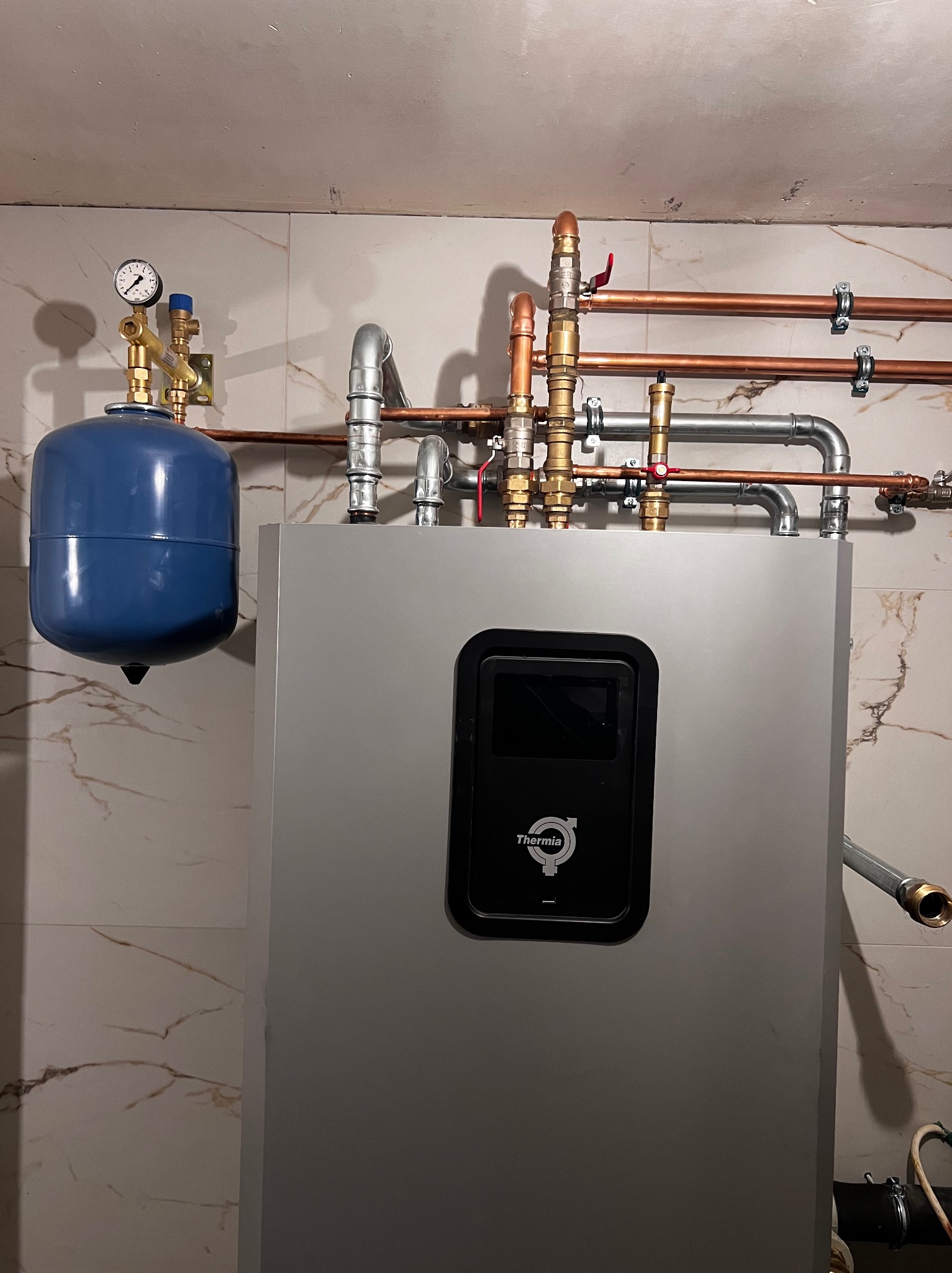 Szary piec Thermia z podłączonymi miedzianymi i stalowymi rurami, niebieskie naczynie wzbiorcze z manometrem, na tle marmurowych płytek ściennych.