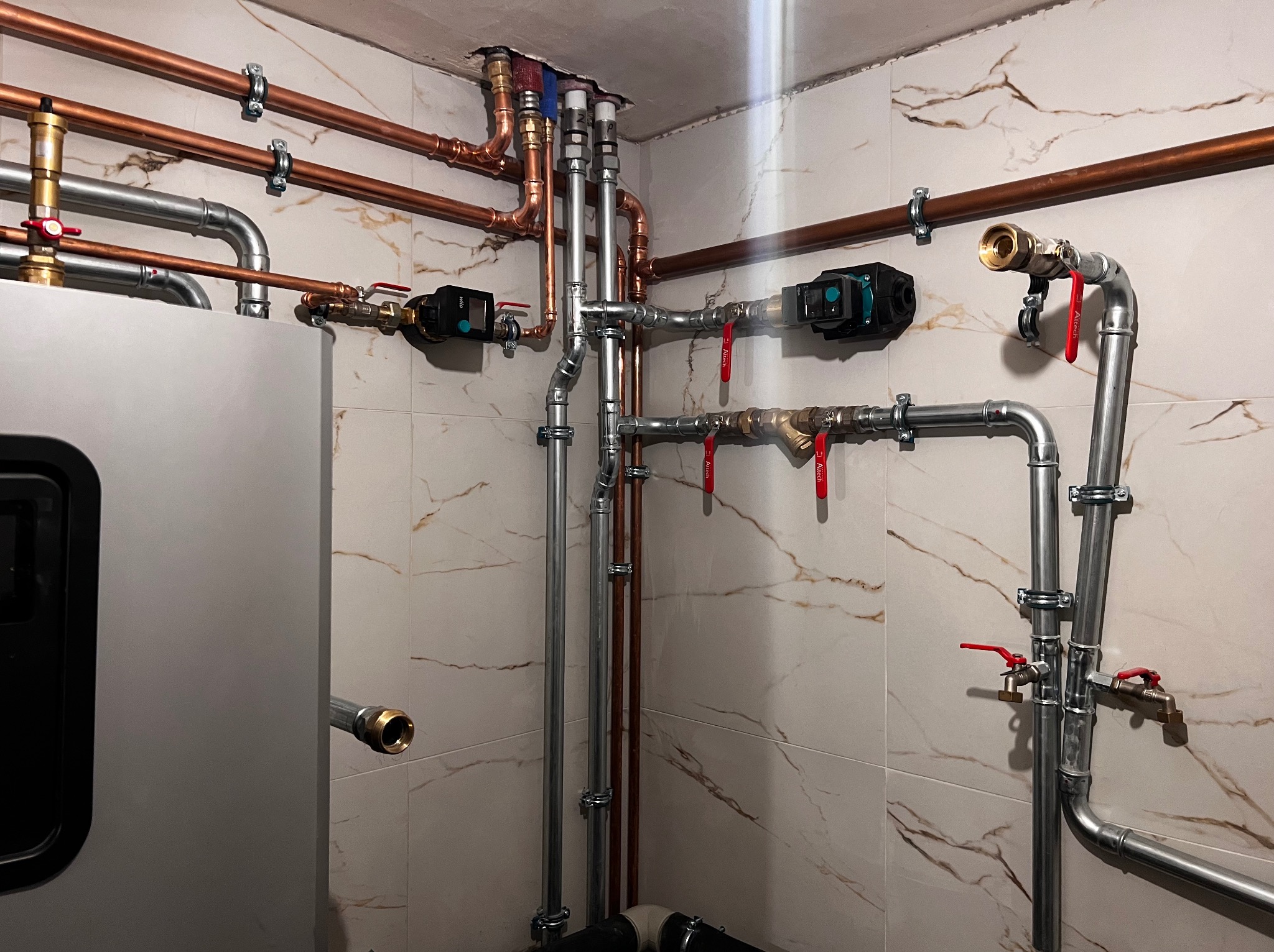 Nowoczesna kotłownia z miedzianymi i stalowymi rurami, pompami obiegowymi i armaturą na tle marmurowych płytek, widoczne elementy instalacji grzewczej i hydraulicznej.
