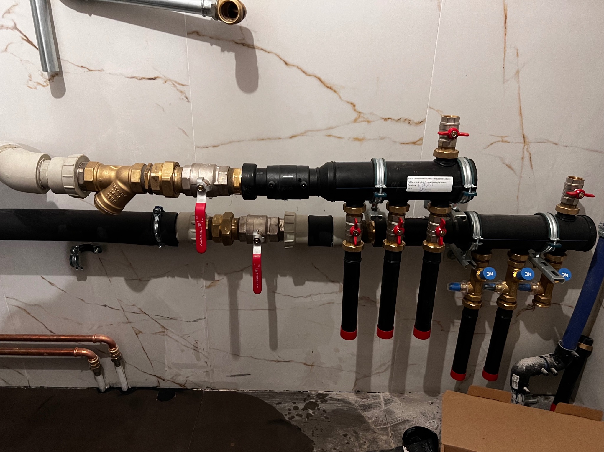 Rozbudowana instalacja wodna z rurami miedzianymi i czarnymi, z licznymi zaworami z czerwonymi i niebieskimi pokrętłami, zamontowana na ścianie z marmurowym wzorem.