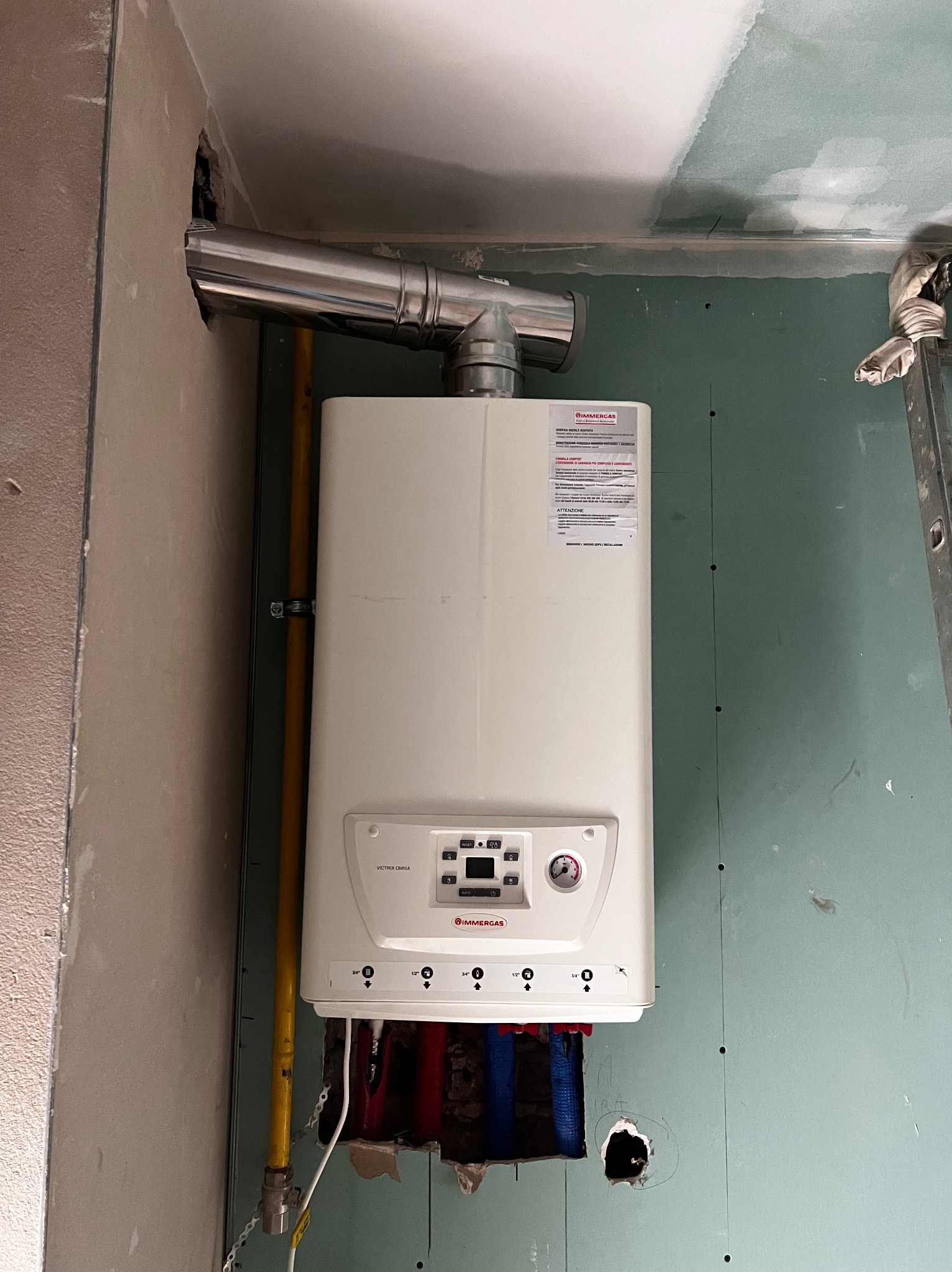 Biały piec gazowy Immergas Victrix Omnia, podłączony do komina i instalacji, widoczny na tle ściany z płyt gipsowo-kartonowych w trakcie montażu.