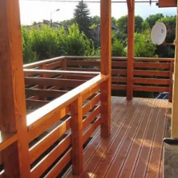 Zadaszony taras z drewnianą podłogą i balustradą w ciepłym odcieniu brązu, widok na ogród i dom z anteną satelitarną w tle, logo firmy widoczne w rogu.