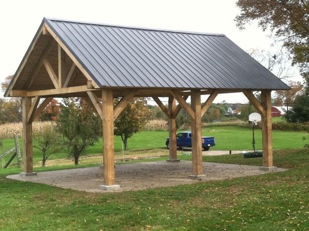 Otwarta konstrukcja z drewnianych belek i metalowym dachem na betonowej podstawie, z widokiem na zielony trawnik, drzewa i zaparkowany niebieski pickup w tle.