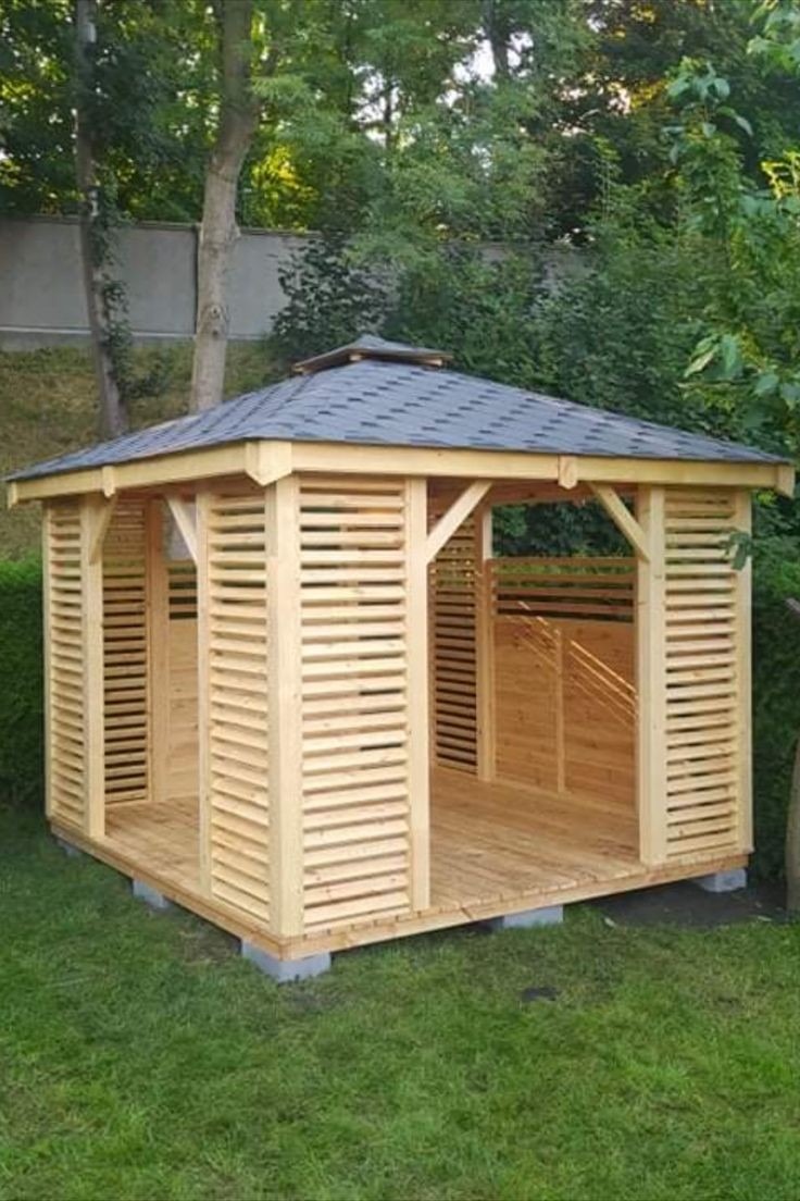 Nowa, jasna konstrukcja z drewna z lamelowymi ścianami i ciemnym dachem z gontów bitumicznych, ustawiona na betonowych bloczkach na trawniku w ogrodzie.