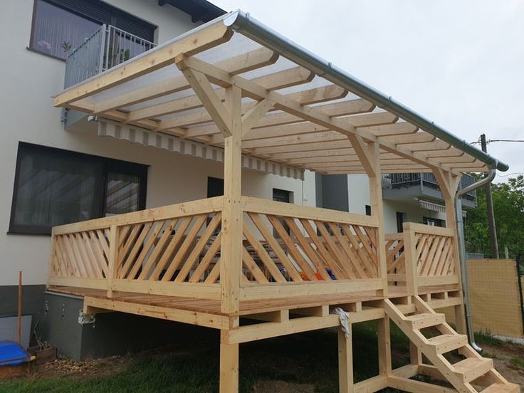 Nowo wybudowany, surowy taras z jasnego drewna z zadaszeniem z przezroczystego tworzywa sztucznego, z balustradą z ukośnych desek i schodami wejściowymi, na tle domu jednorodzinnego.