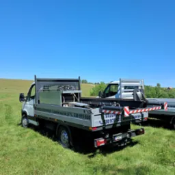 Dwa samochody dostawcze z zabudową do wiercenia studni głębinowych na trawiastym terenie, widoczne numery telefonów i adresy stron internetowych na zabudowie. Błękitne niebo w tle.