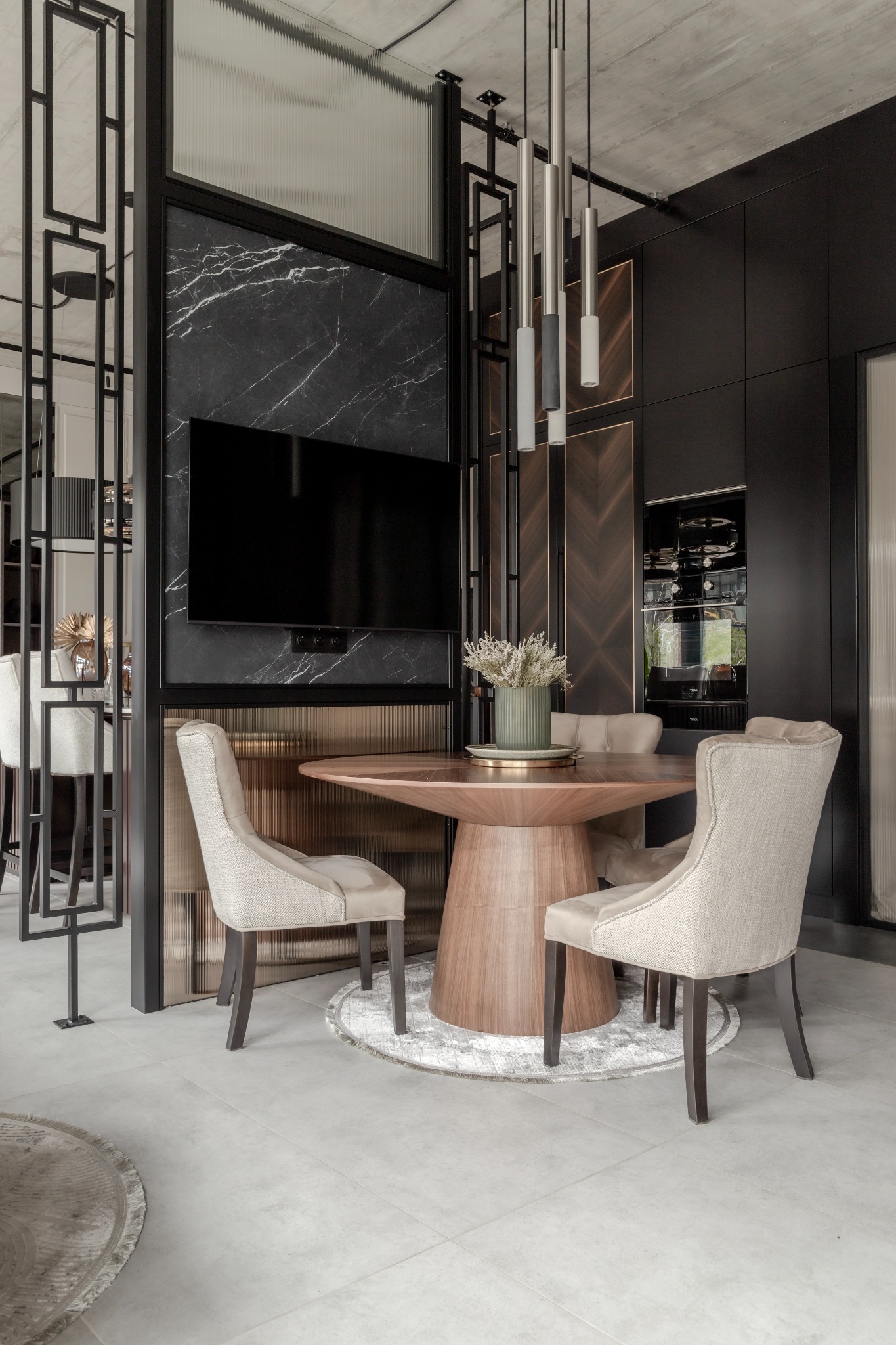 Nowoczesna jadalnia z okrągłym drewnianym stołem, tapicerowanymi krzesłami i dekoracyjnymi lampami wiszącymi na tle ściany z czarnego marmuru z zamontowanym telewizorem.