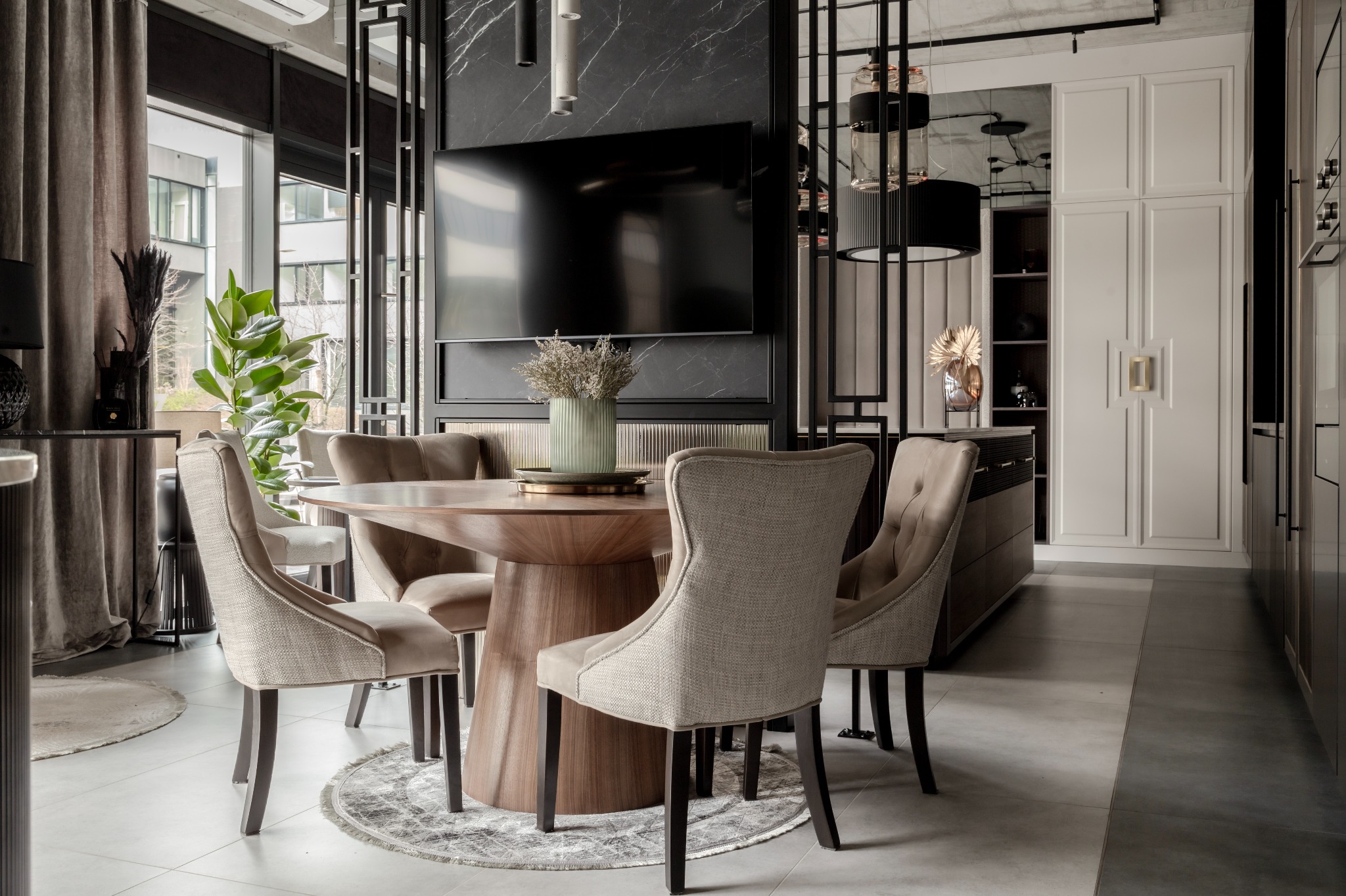 Nowoczesna jadalnia z okrągłym drewnianym stołem na jednej nodze, otoczonym przez tapicerowane krzesła w beżowej kolorystyce, na tle marmurowej ściany z telewizorem i zabudowy meblowej.