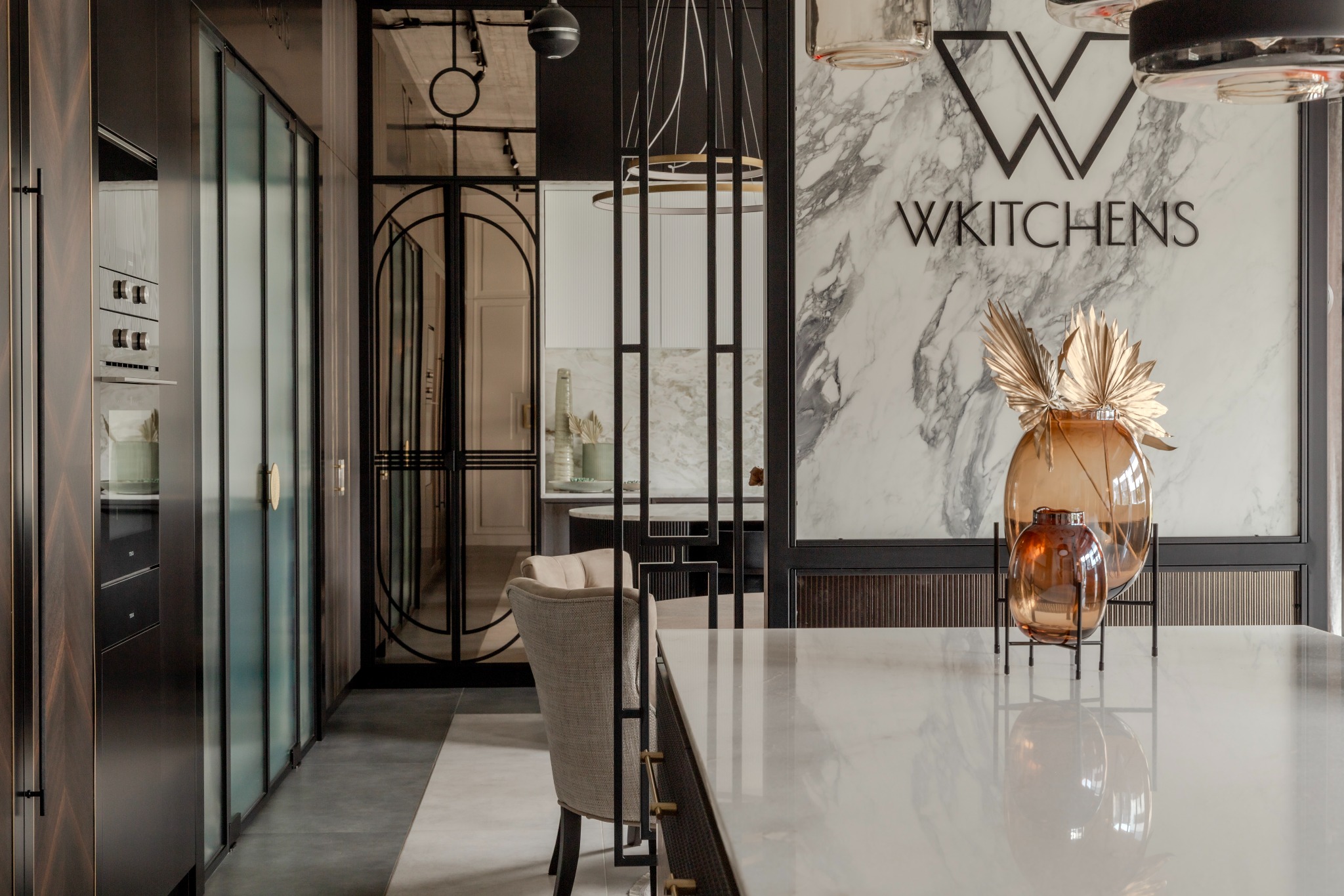 Elegancka kuchnia z marmurową wyspą i ścianą akcentową, szklane fronty szafek, dekoracyjne wazony z suszonymi roślinami, logo 'W KITCHENS' na marmurze.