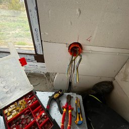Elektro-Kwiatek - Pomarańczowa puszka instalacyjna z wystającymi kablami w surowym wnętrzu, obok skrzynka z konektorami, obcęgi, nóż i fragment spodni roboczych.