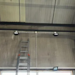 Dwa reflektory LED zamontowane na suficie hali, połączone rurką instalacyjną, drabina aluminiowa oparta o ścianę, widoczne okno w dolnej części kadru.