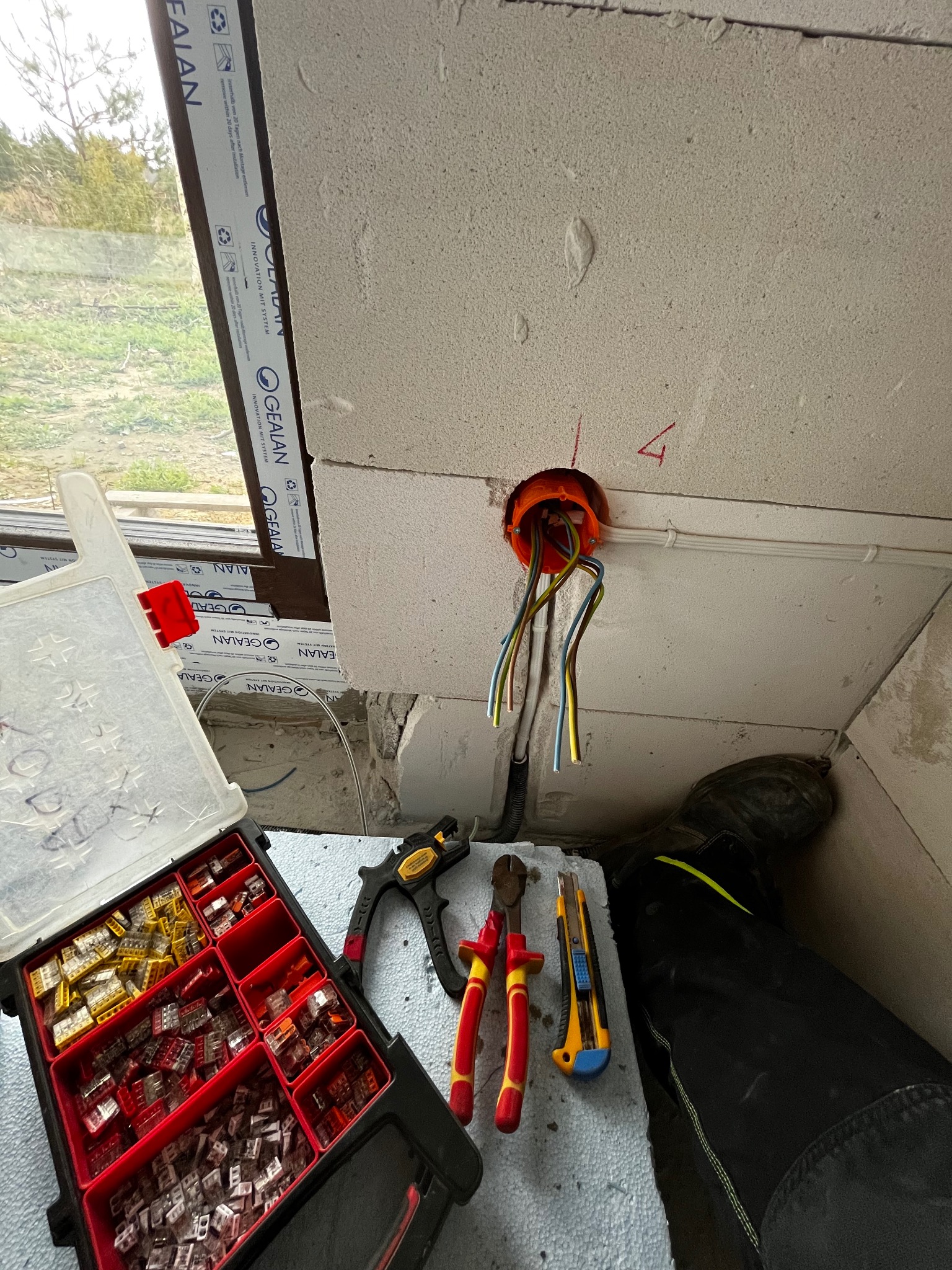 Pomarańczowa puszka instalacyjna z wystającymi kablami w surowym wnętrzu, obok skrzynka z konektorami, obcęgi, nóż i fragment spodni roboczych.