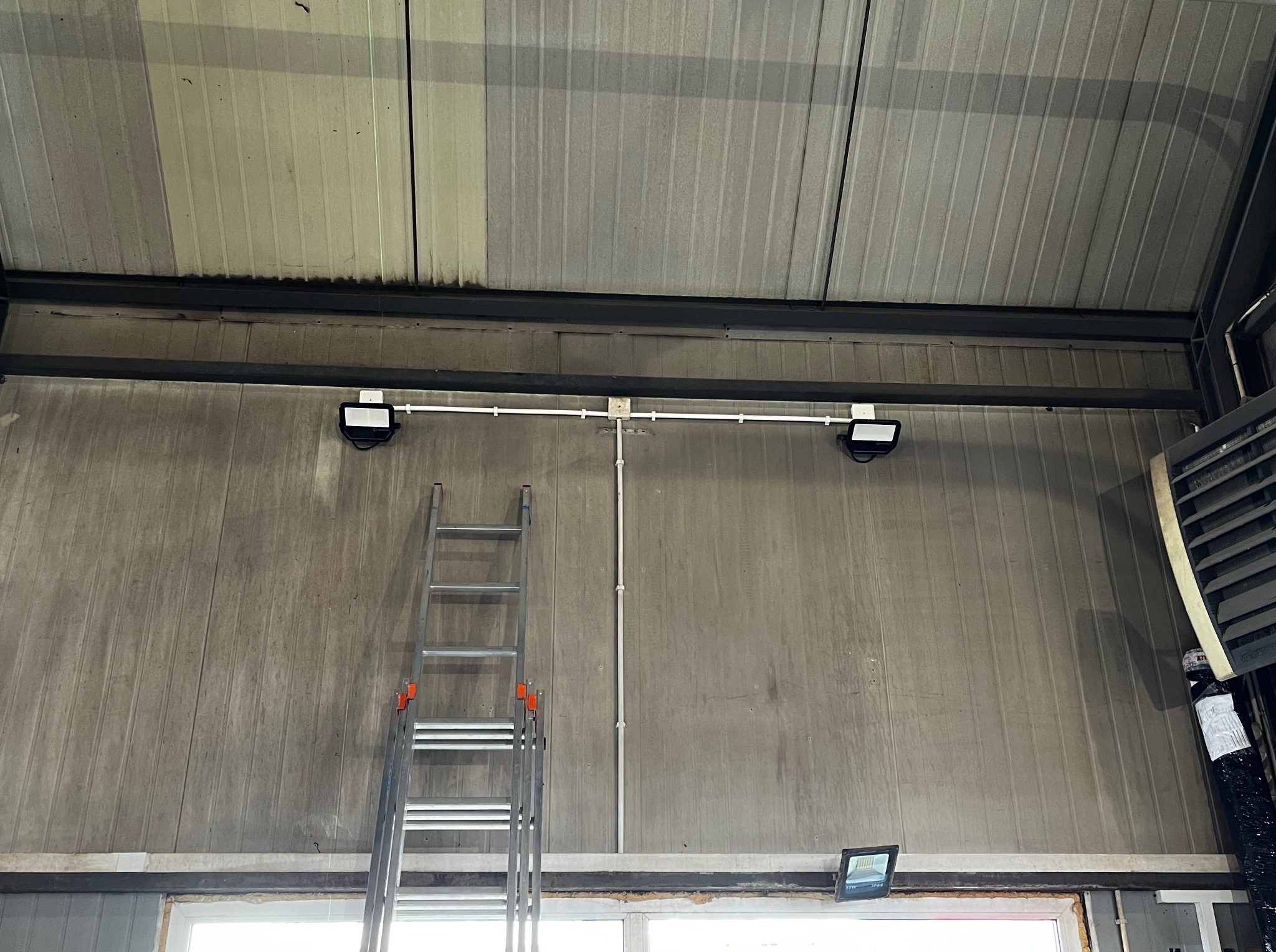 Dwa reflektory LED zamontowane na suficie hali, połączone rurką instalacyjną, drabina aluminiowa oparta o ścianę, widoczne okno w dolnej części kadru.