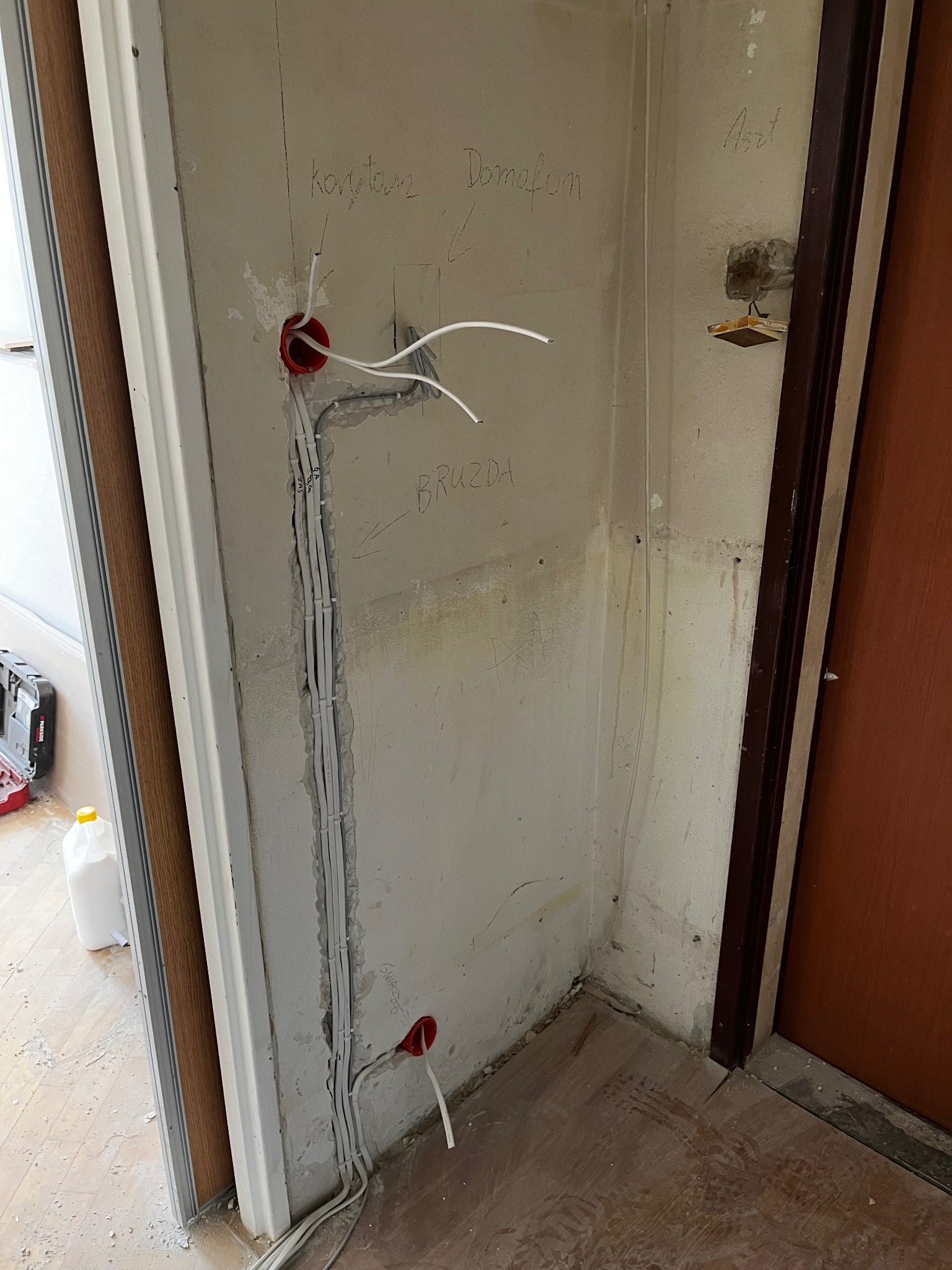 Ściana z wyciętą bruzdą pod przewody elektryczne, widoczne kable w peszlu, puszki instalacyjne, oraz odręczne notatki na ścianie. Podłoga z panelami.