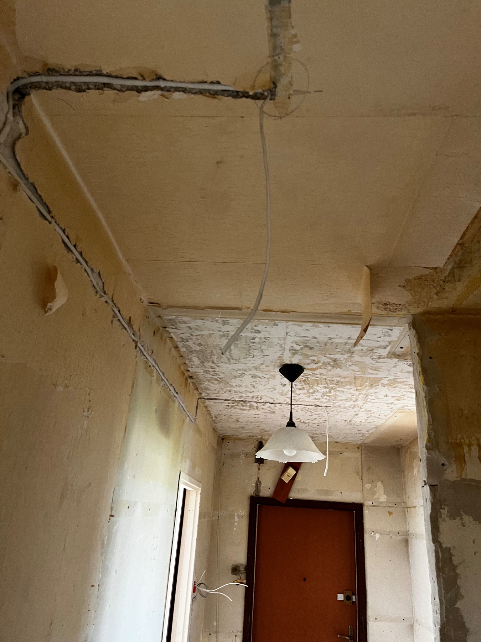 Wnętrze w trakcie remontu: widoczne przewody elektryczne poprowadzone wzdłuż sufitu i ściany, wisząca lampa z żarówką, ściany w stanie surowym z resztkami tapet i farby.