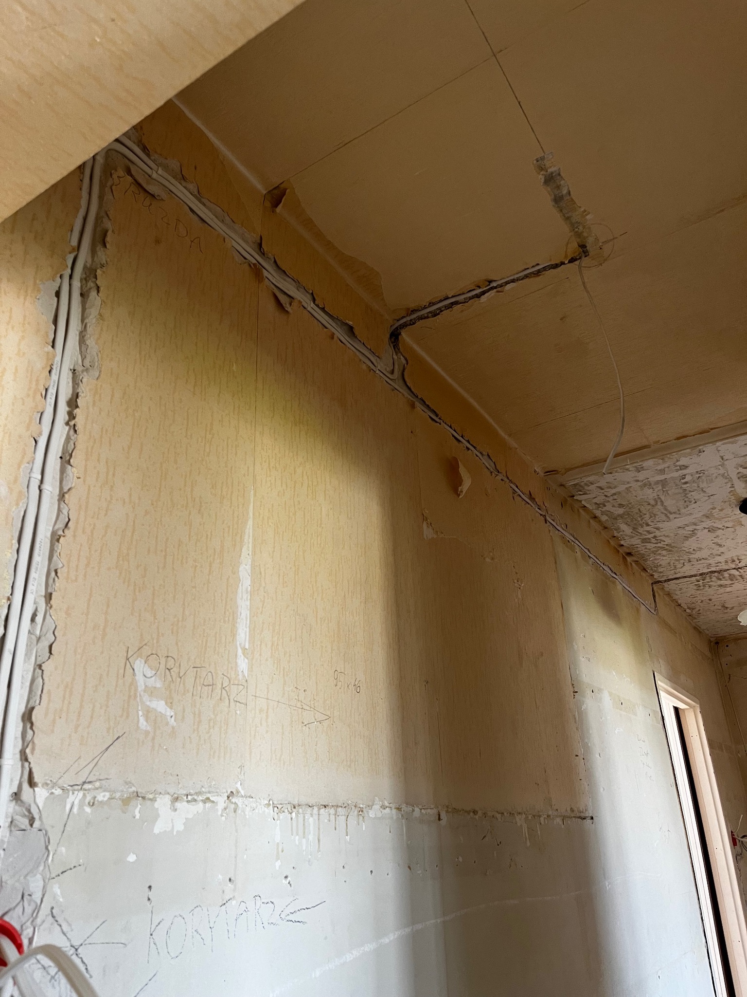 Białe przewody elektryczne ułożone w bruzdach w ścianie i suficie podczas remontu, widoczne ślady po kuciu i napisy 'Korytarz' na ścianie.