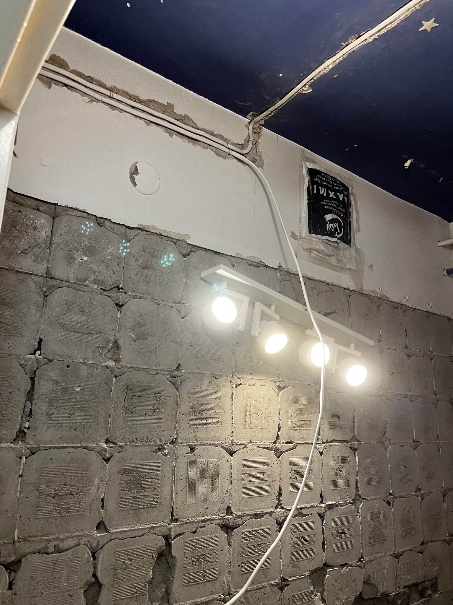 Biała listwa z pięcioma reflektorami zamontowana na surowej, betonowej ścianie z widocznymi instalacjami elektrycznymi i fragmentem niebieskiego sufitu ze sztucznymi gwiazdami.