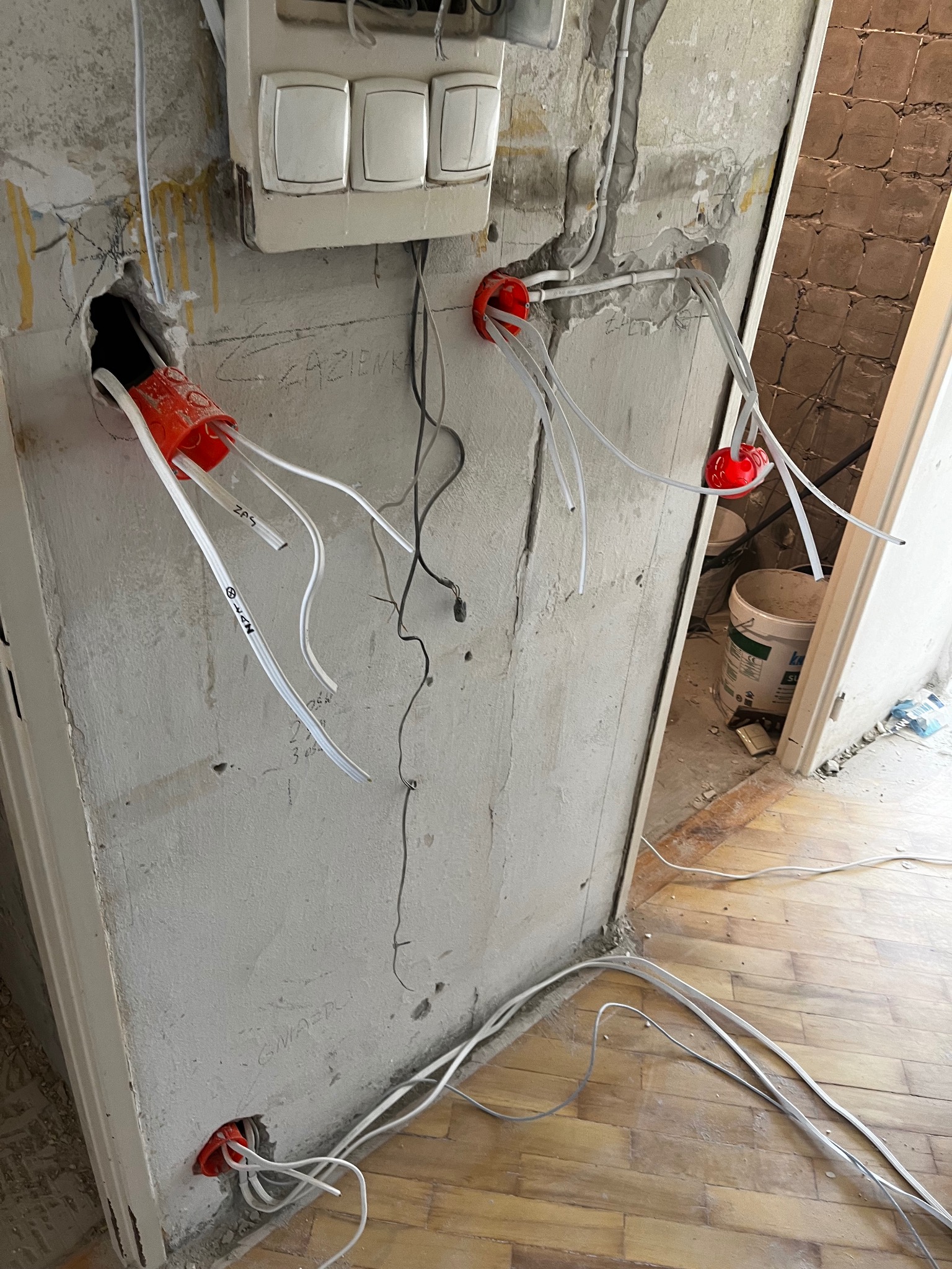Wewnętrzne okablowanie elektryczne w trakcie montażu w surowej ścianie z widocznymi puszkami instalacyjnymi i przewodami z oznaczeniami, w tle fragment drewnianej podłogi i otwarte drzwi.
