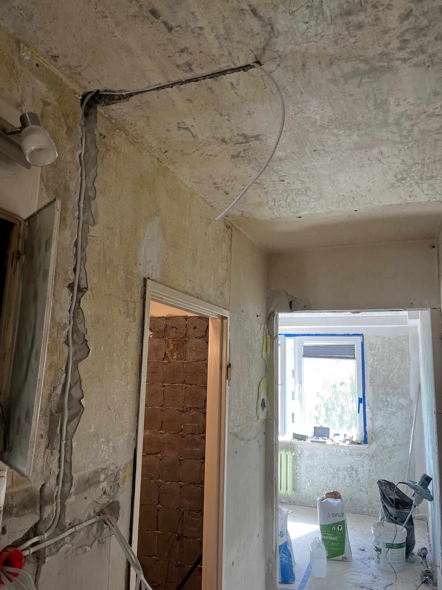 Wnętrze w trakcie remontu: widoczne kable elektryczne prowadzone po ścianie i suficie, odsłonięte fragmenty muru, worki z zaprawą i narzędzia budowlane.