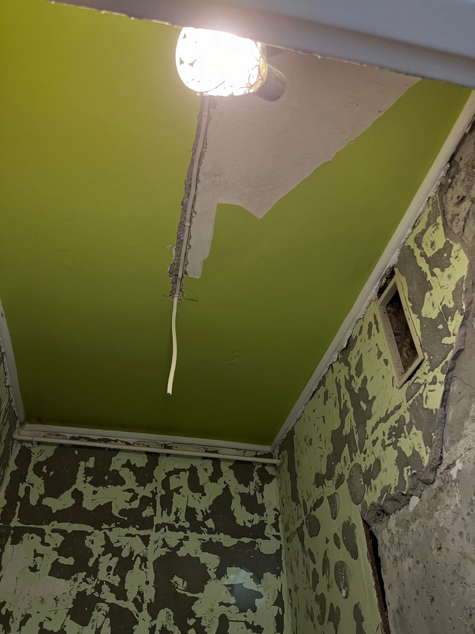 Niedokończone wnętrze: surowy tynk na ścianach, fragment pomalowanego na zielono sufitu z wiszącym przewodem elektrycznym i żarówką.