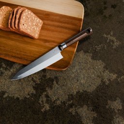 Kamil Macieja - Profesjonalny nóż szefa kuchni z drewnianą rękojeścią leży na drewnianej desce do krojenia obok pokrojonego chleba na tle surowej kamiennej powierzchni.