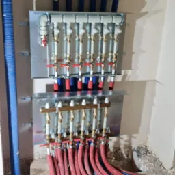 Rozdzielacz instalacji grzewczej podłączony do rur czerwonych i niebieskich, zamontowany na ścianie w surowym stanie.