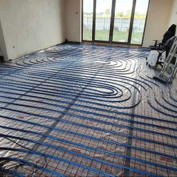 INSTALTOR, instaltor.pl - Niebieskie rury ogrzewania podłogowego ułożone spiralnie na folii refleksyjnej z czerwoną siatką w surowym wnętrzu, widoczna drabina i okno tarasowe.