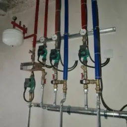 Rozbudowana instalacja hydrauliczna i grzewcza z rurami w kolorach czerwonym i niebieskim, zaworami, pompami obiegowymi i manometrami, zamontowana pod sufitem w surowym stanie.