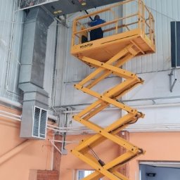 INSTALTOR, instaltor.pl - Pracownik na podnośniku nożycowym instaluje system wentylacyjny w hali, widoczne kanały wentylacyjne i kratki wentylacyjne pod sufitem.