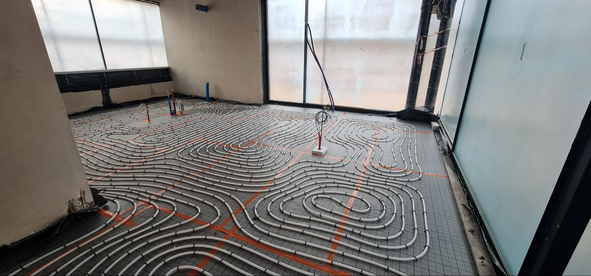 Rozłożone rury ogrzewania podłogowego na czarnej macie z pomarańczową siatką w surowym wnętrzu z dużymi oknami, widoczne pionowe rury instalacji.